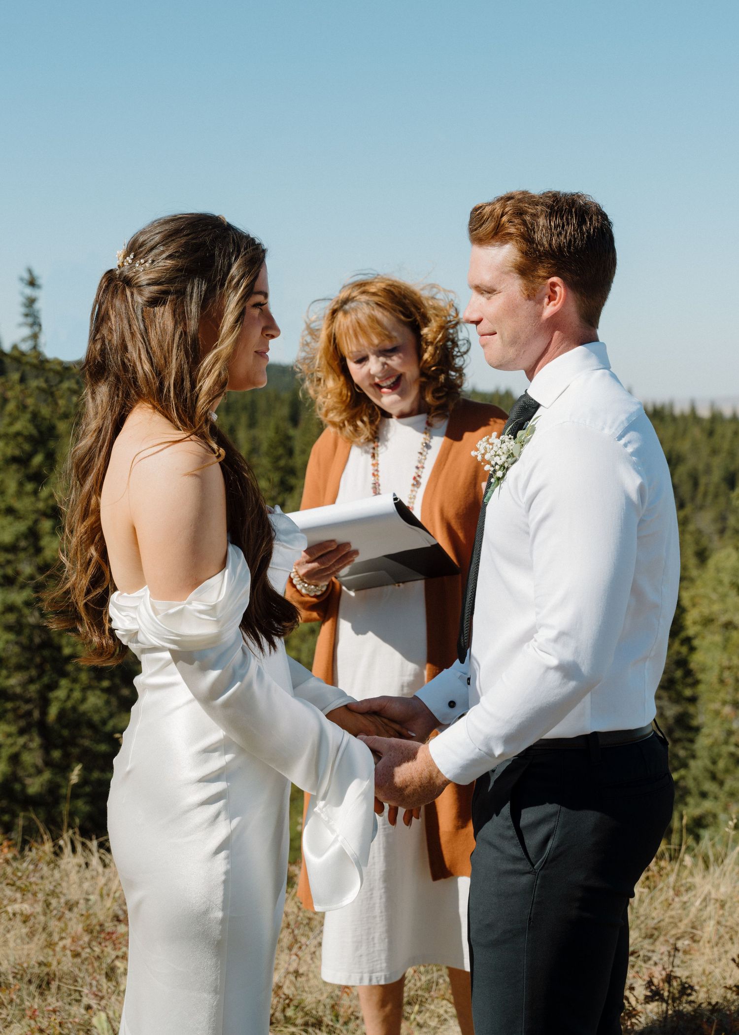 Bride and groom exchange vows outdoors with officiant and pine tree backdrop under blue sky.