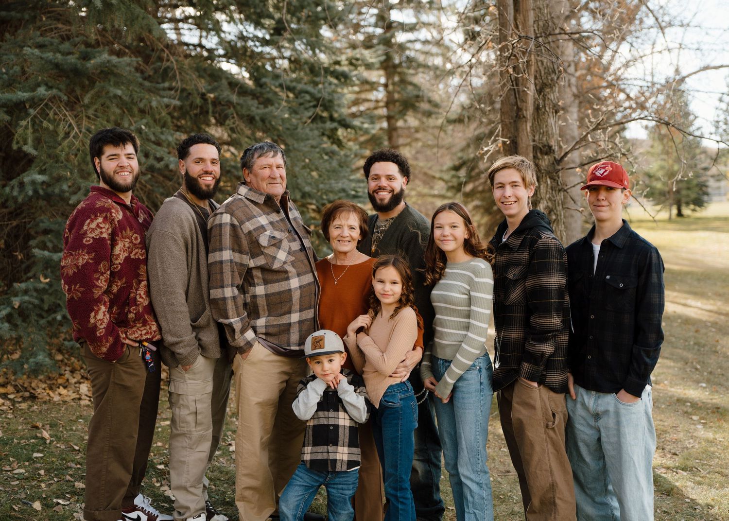 Extended family group of nine posing outdoors near pine trees in casual autumn attire.