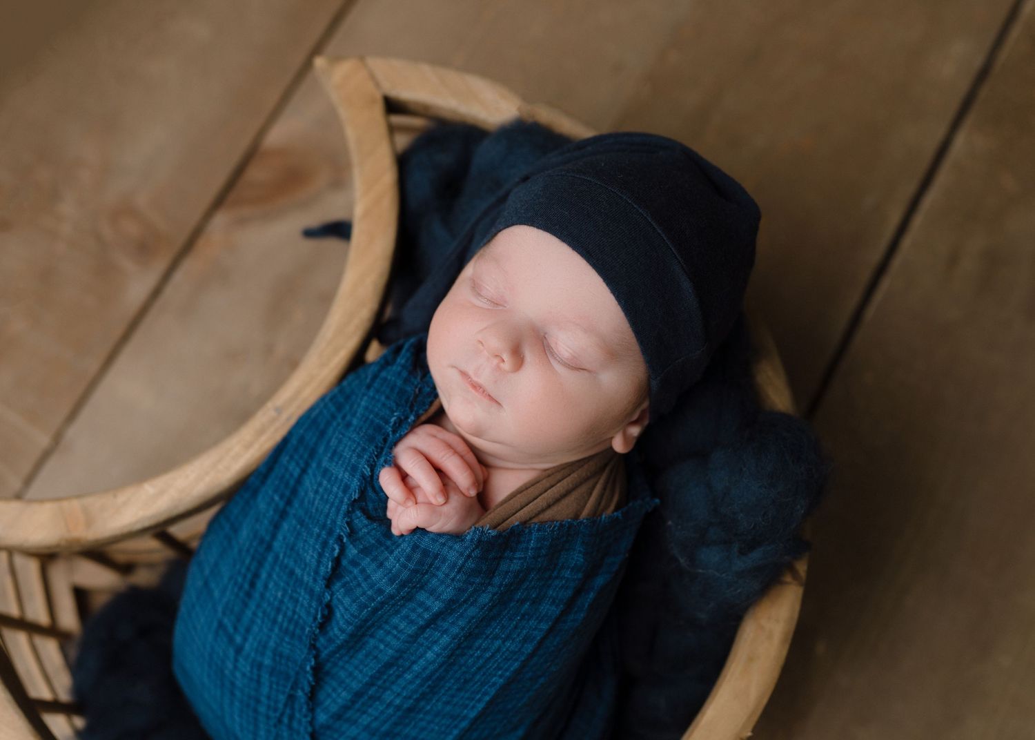 Sleeping newborn baby wrapped in navy blue swaddle with matching knit hat resting in a wicker basket.