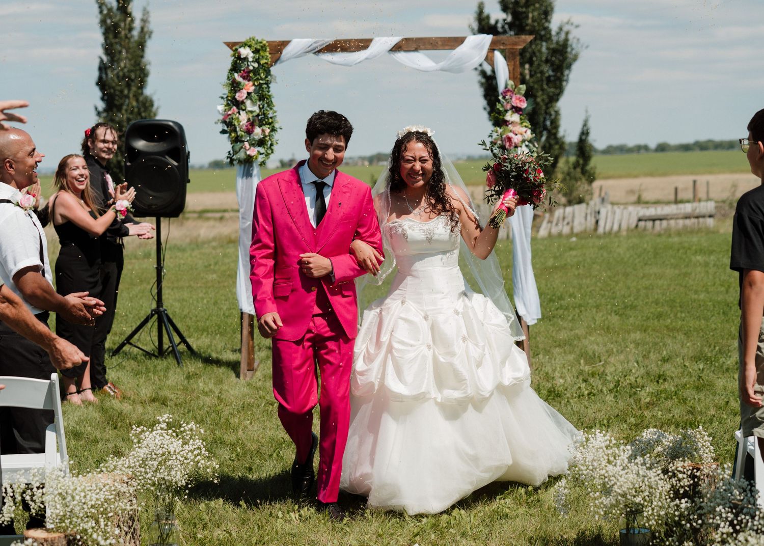 Bride in white gown and groom in pink suit walk down outdoor wedding aisle as guests cheer.