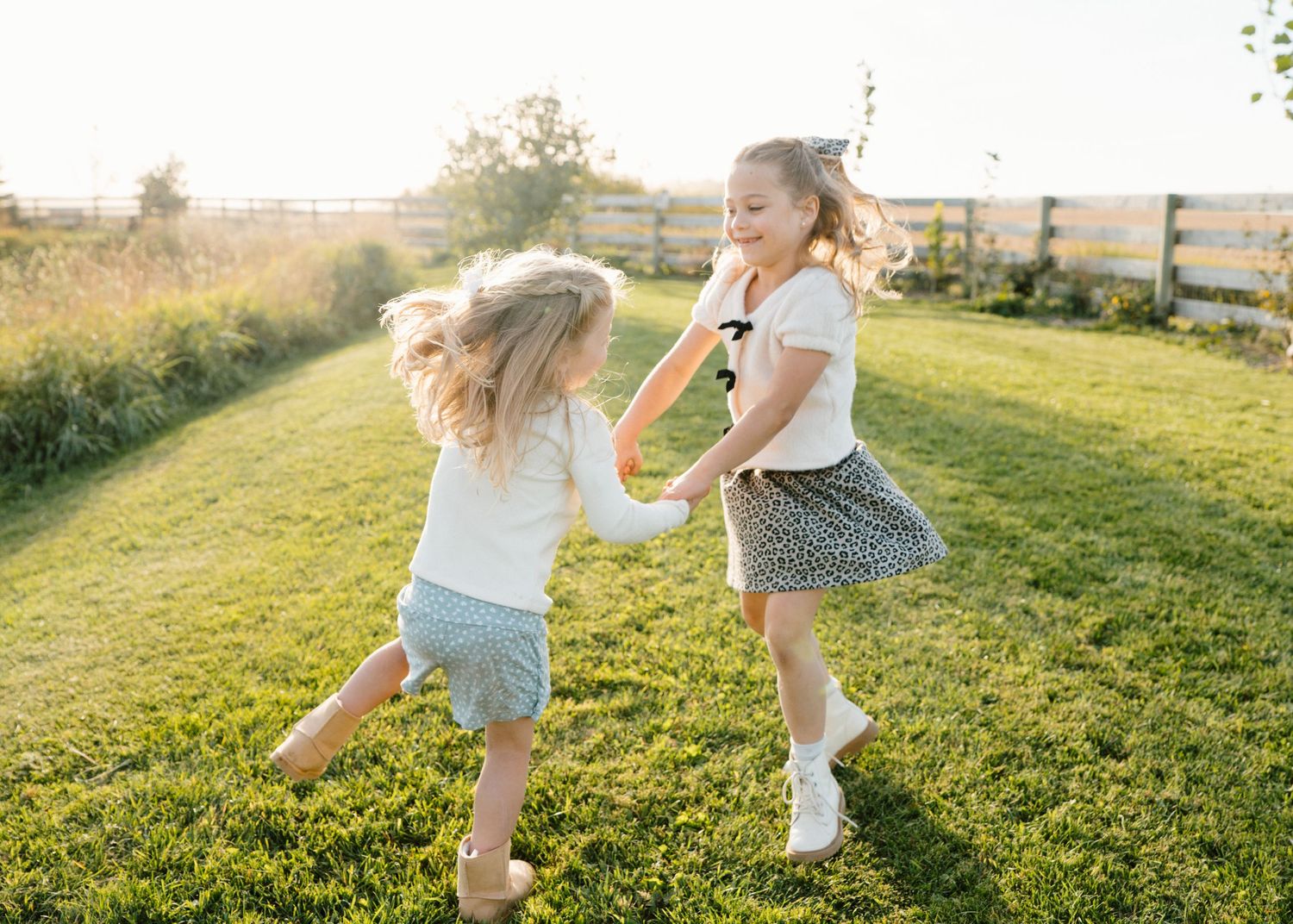 Two young girls holding hands and spinning together on a sunny green lawn near a wooden fence.