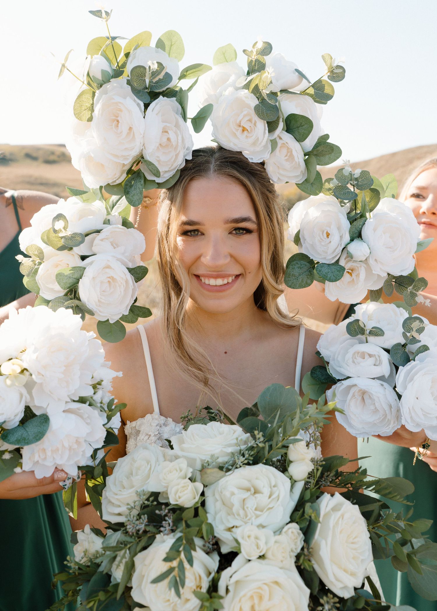 Smiling bride framed by white roses and eucalyptus bouquet held by bridesmaids in emerald dresses.