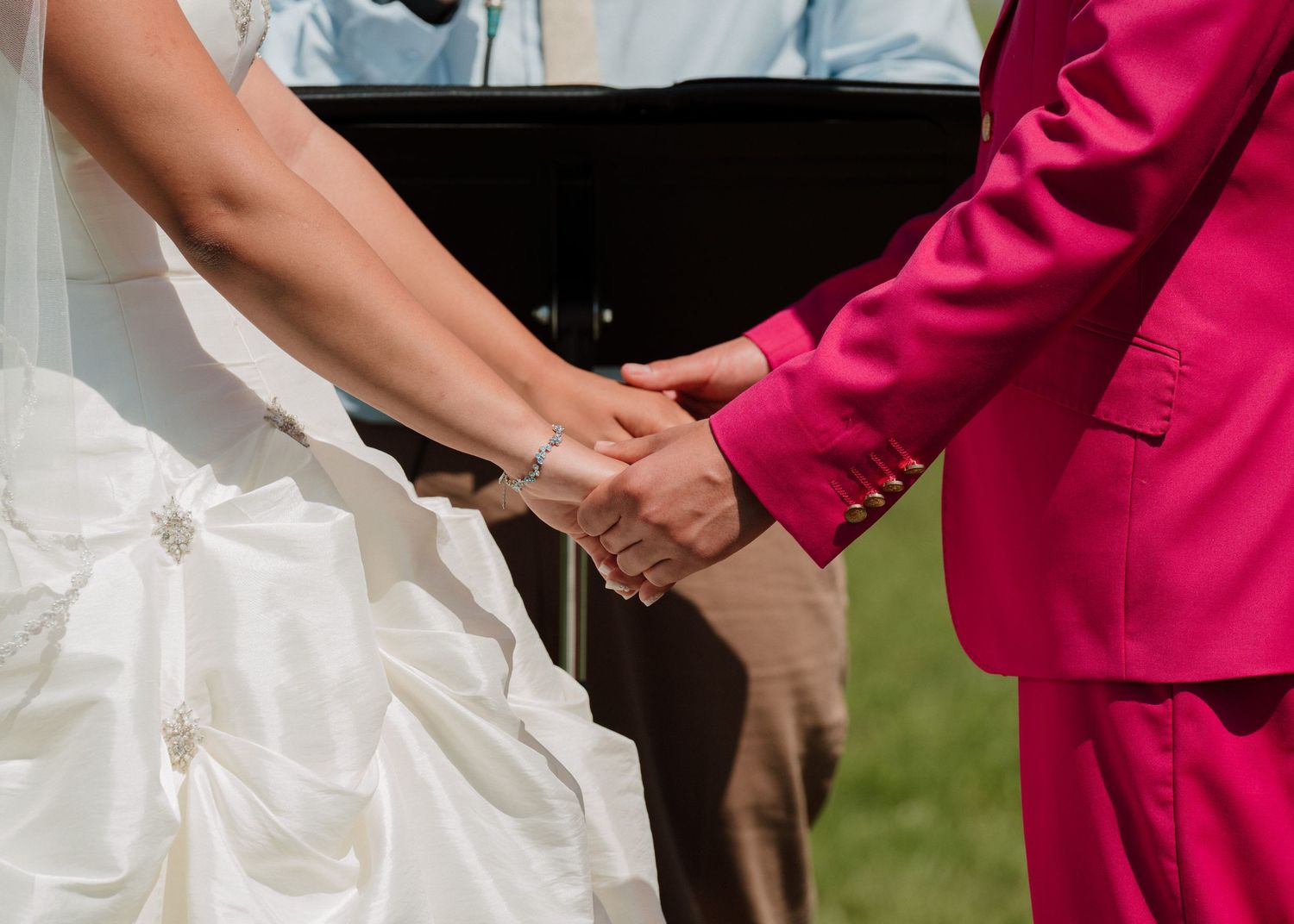 Bride and groom hold hands during outdoor wedding ceremony, groom wearing a bold pink suit.