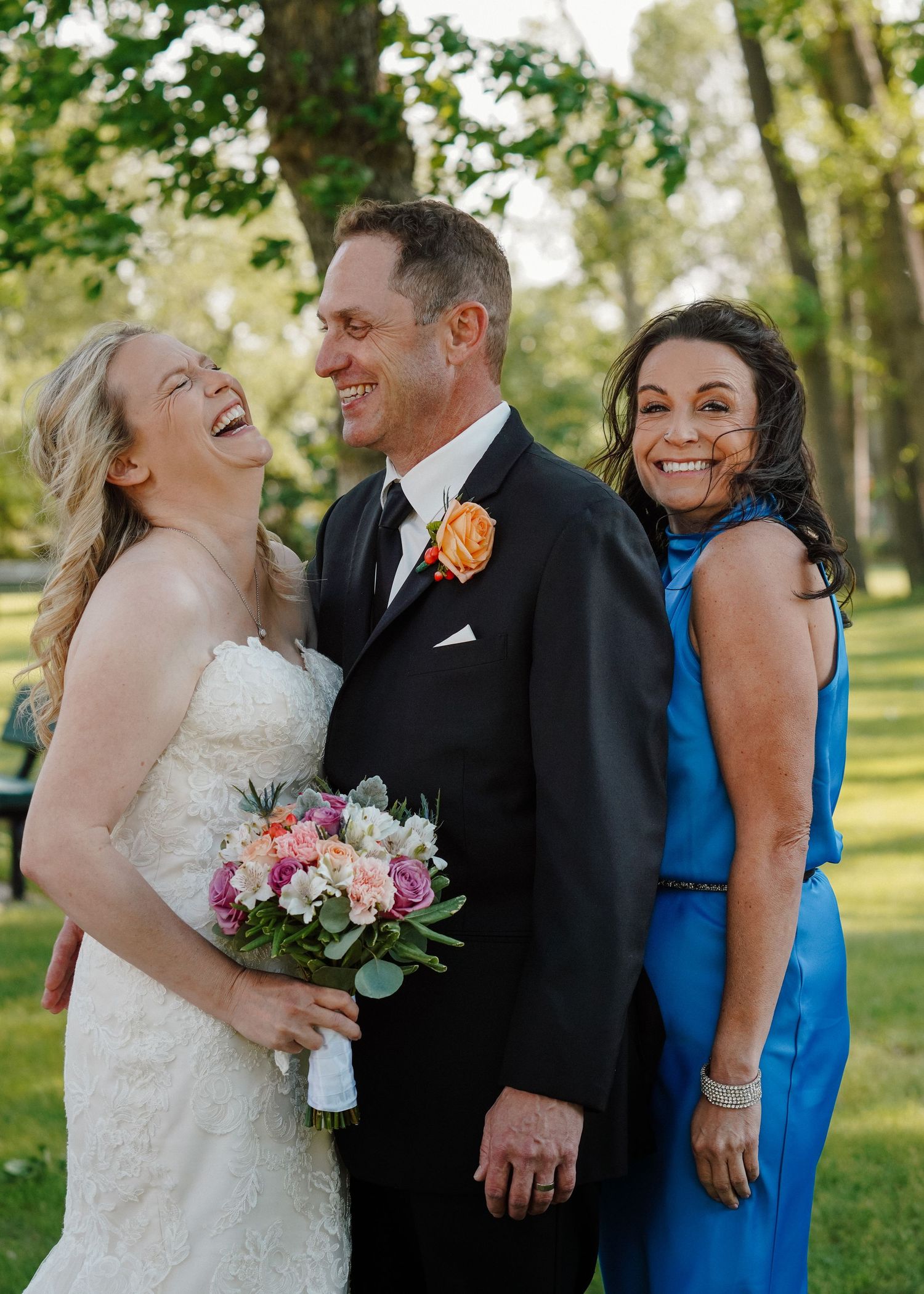 Bride and groom laugh joyfully with a woman in blue dress during outdoor wedding party photos.