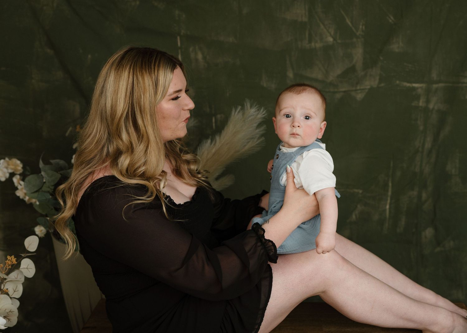 Blonde woman holding a young baby against a dark green studio backdrop.