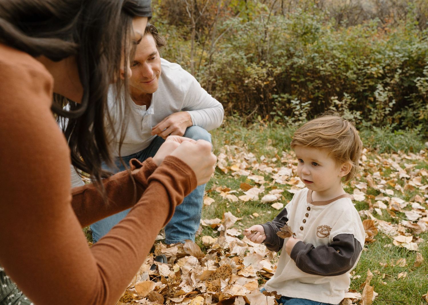 Young parents engage with their curious toddler son while playing in a pile of autumn leaves.