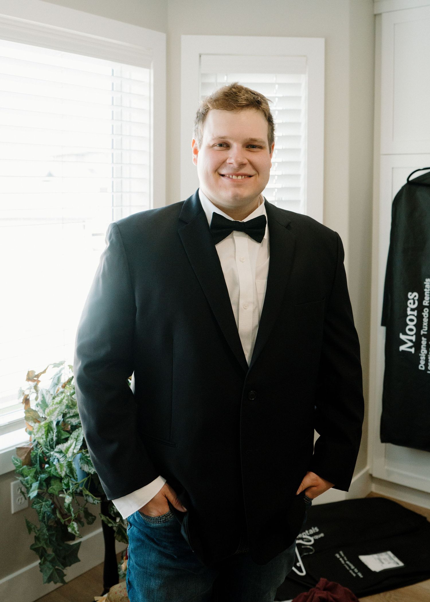 Smiling groom in black tuxedo and bow tie standing by window before wedding ceremony.