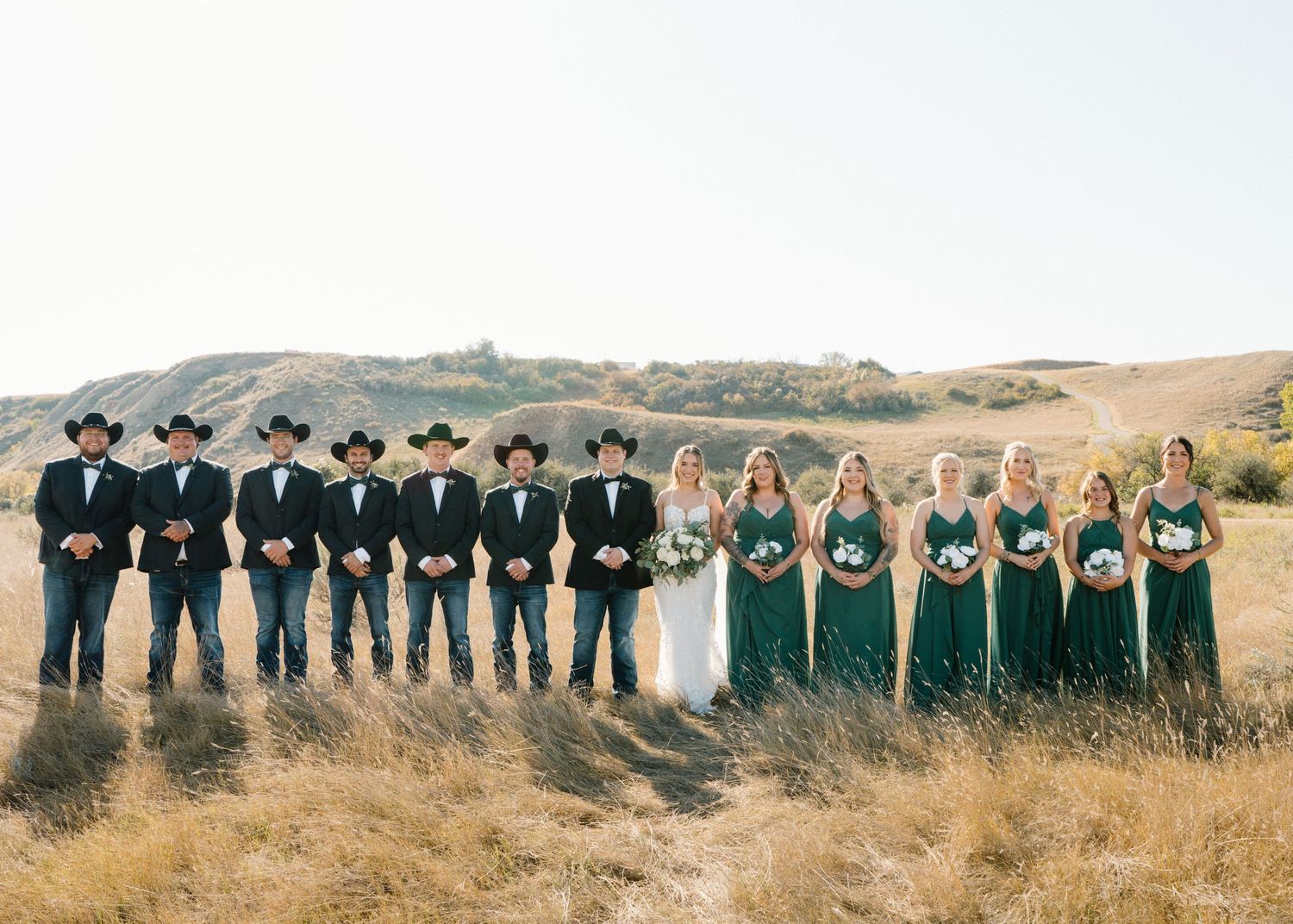 Western wedding party split into two groups in golden field, bridesmaids in emerald, groomsmen in black.