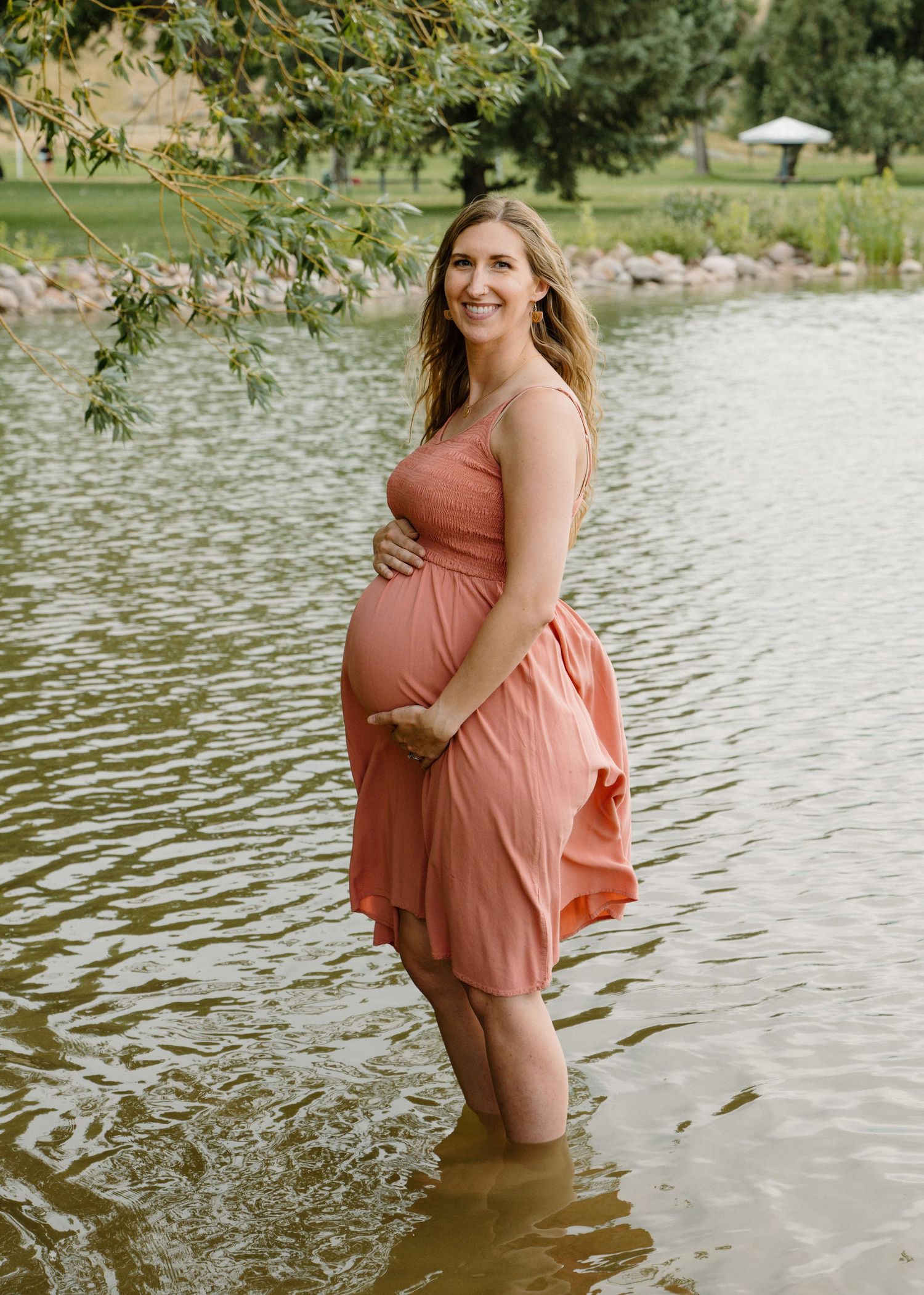 Smiling pregnant woman in a coral dress standing in a pond with green trees behind.