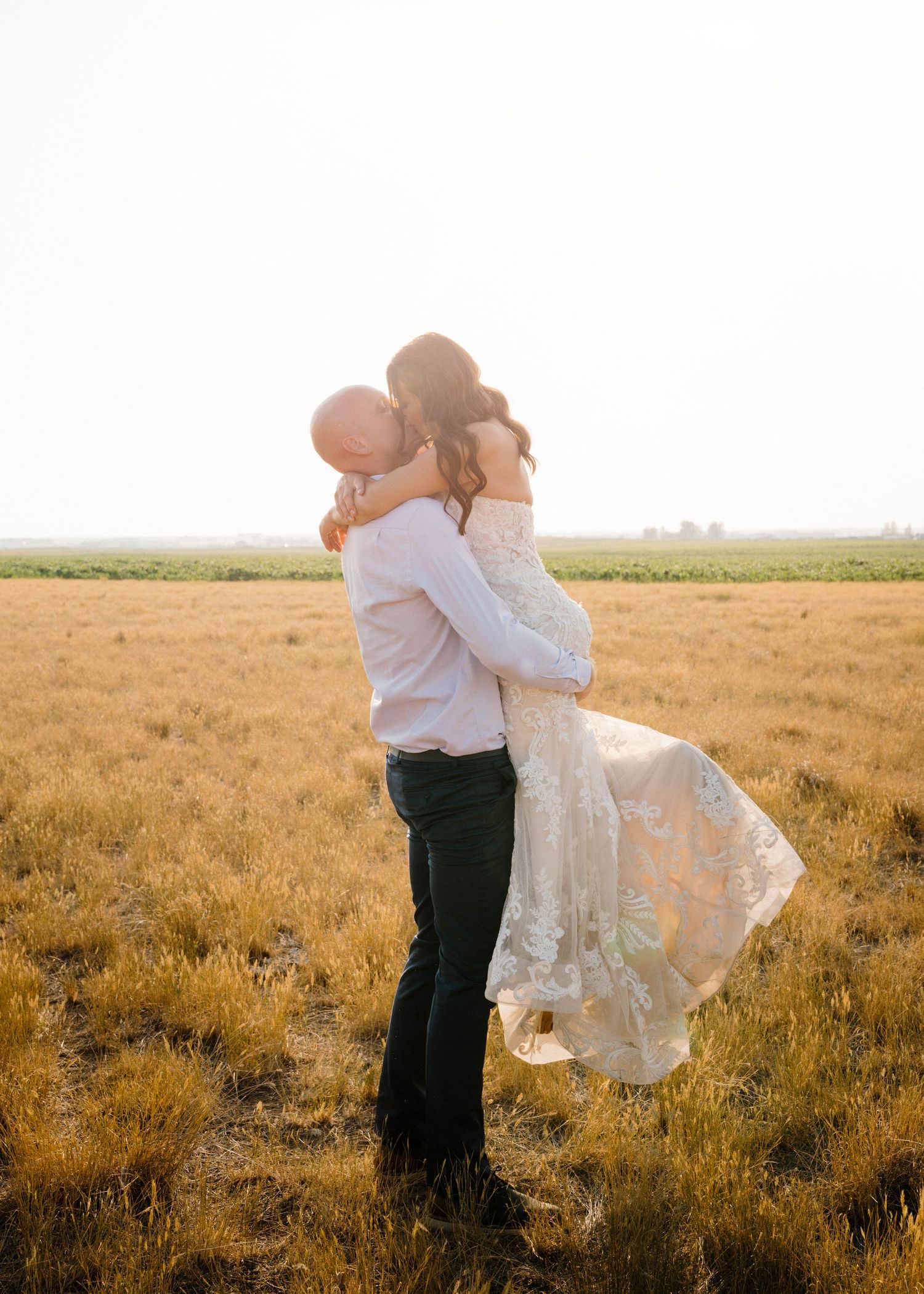 Groom lifts bride in a romantic embrace in a sunlit golden field during outdoor wedding photos.
