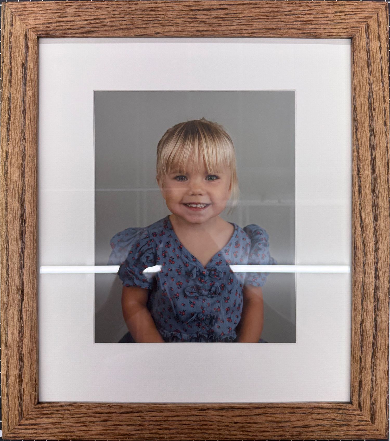 Framed portrait photo of a smiling young blonde girl wearing a blue floral dress, displayed in a wooden oak frame with white matting.