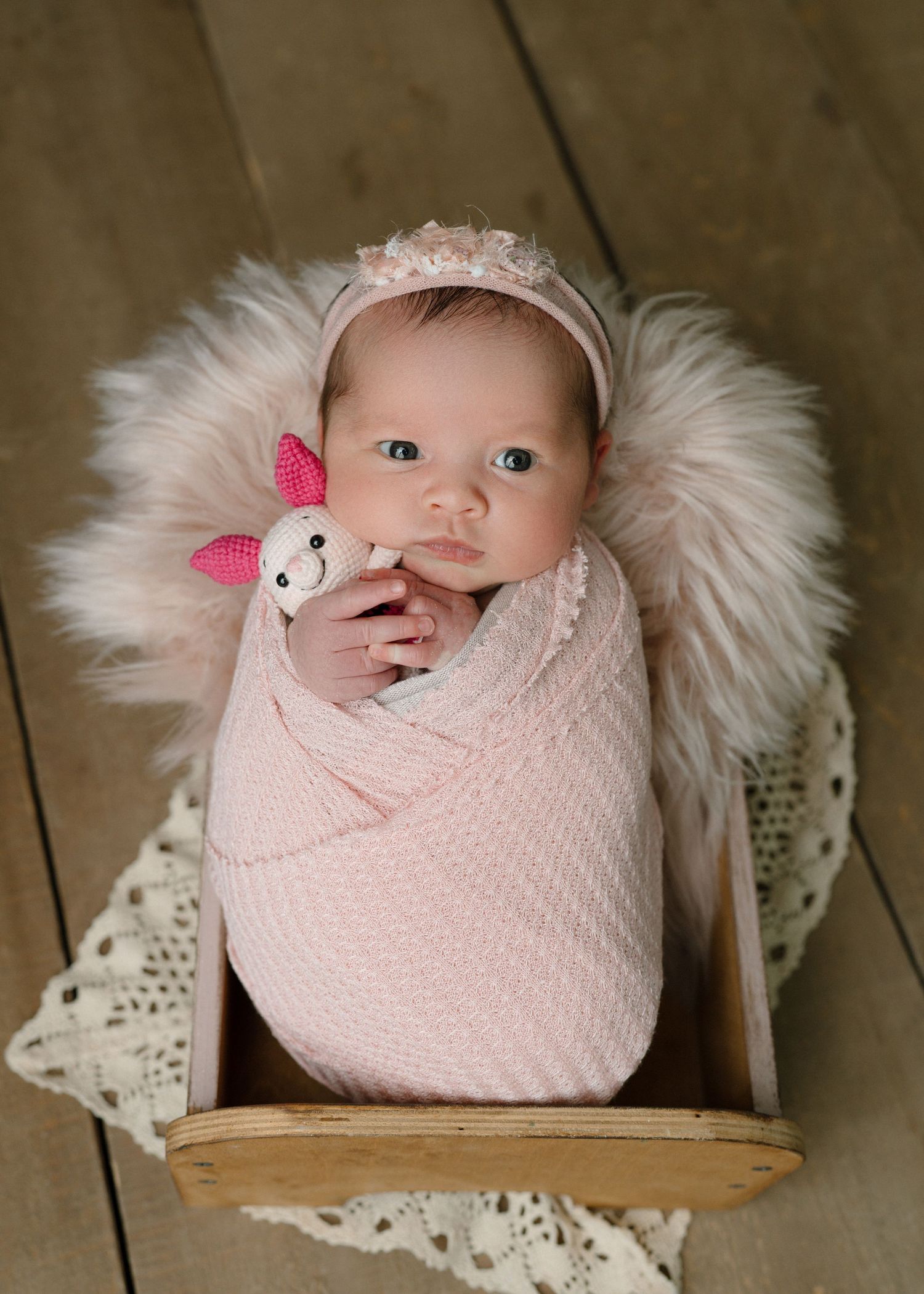 Newborn baby girl swaddled in pink wrap with floral headband, holding a bunny rattle in a wooden crate with fur.