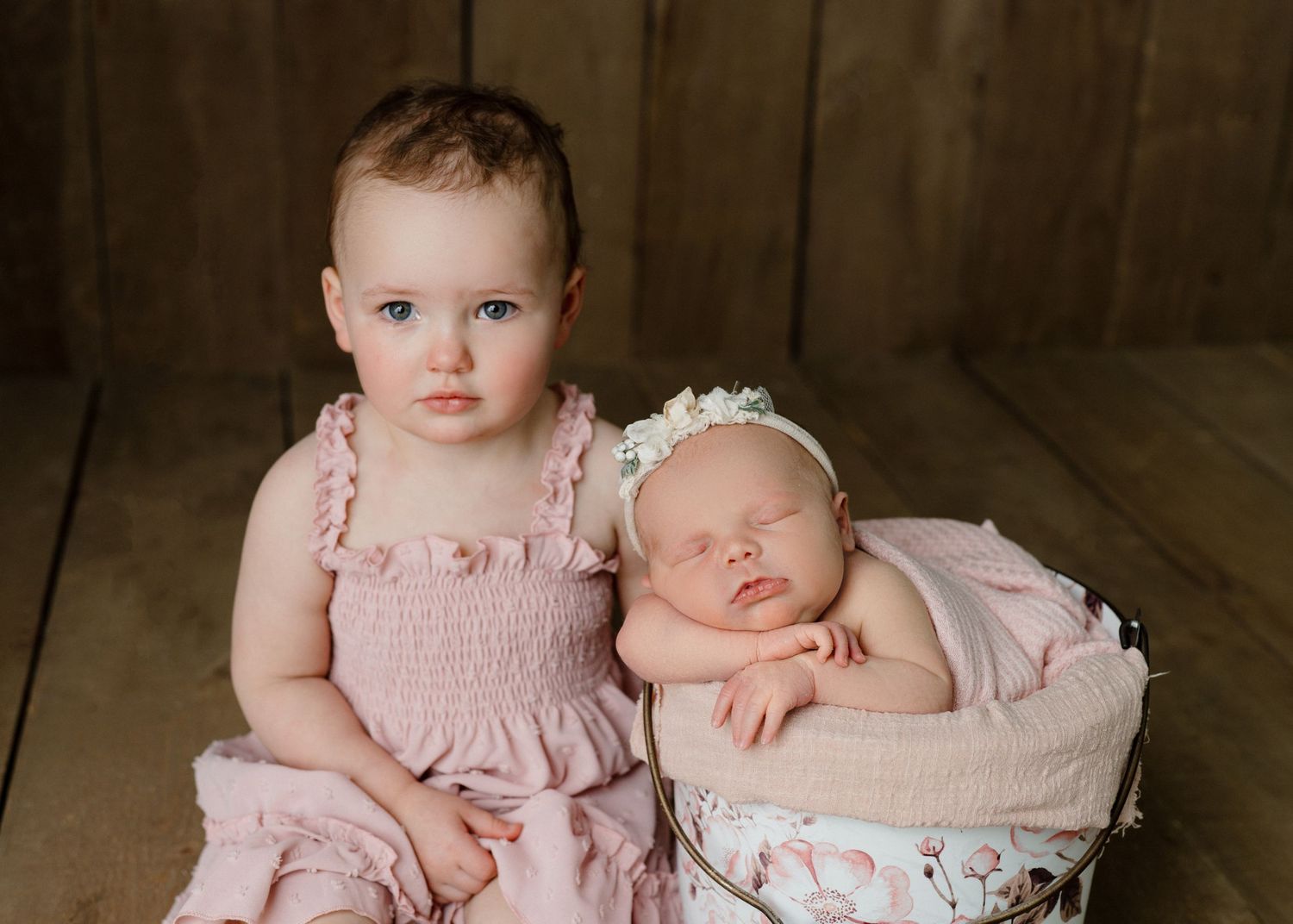 Toddler girl in pink dress sits beside sleeping newborn baby in a white basket for a sibling portrait.