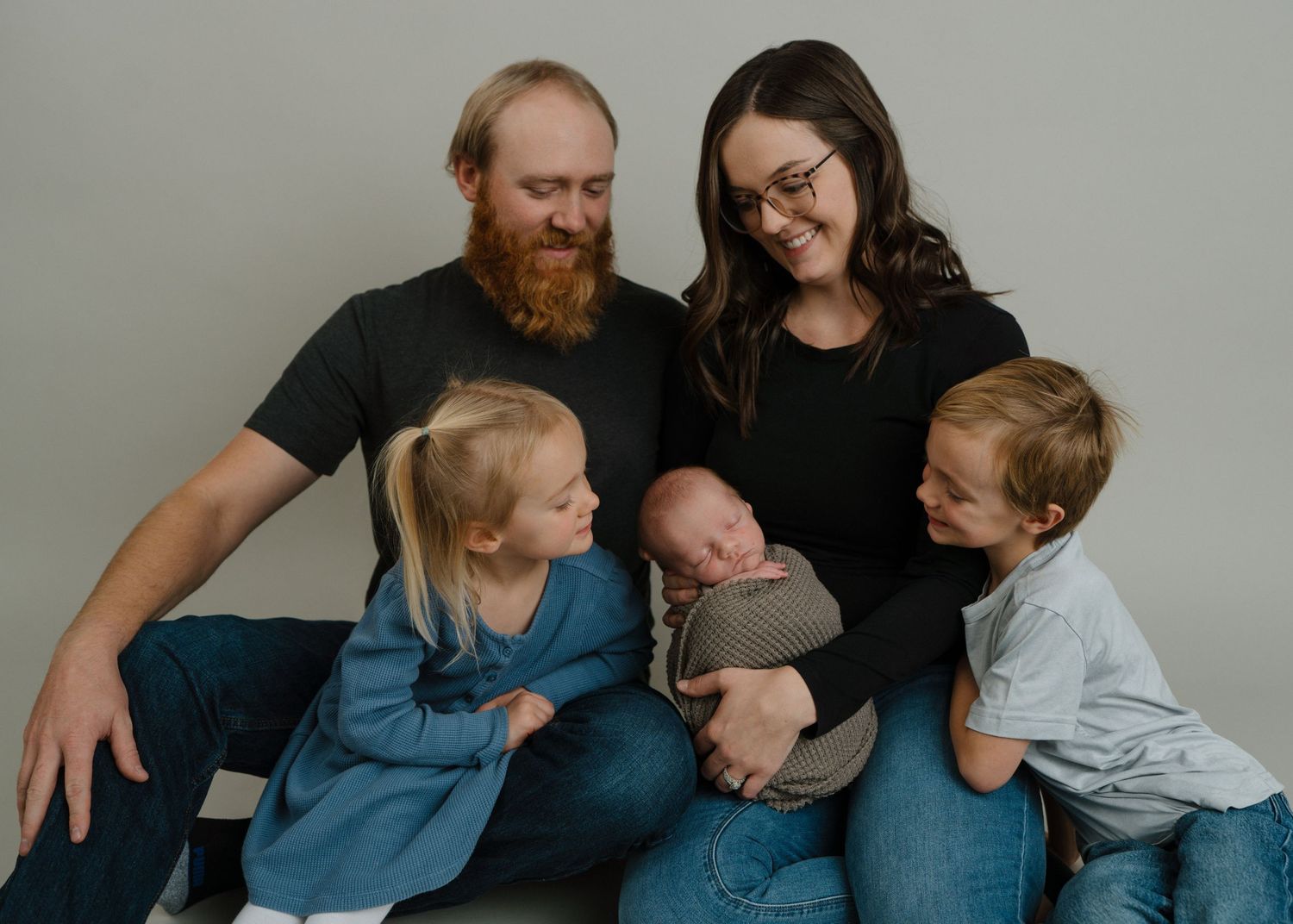 Happy family of five with newborn baby, two older children, mom and bearded dad on grey backdrop.