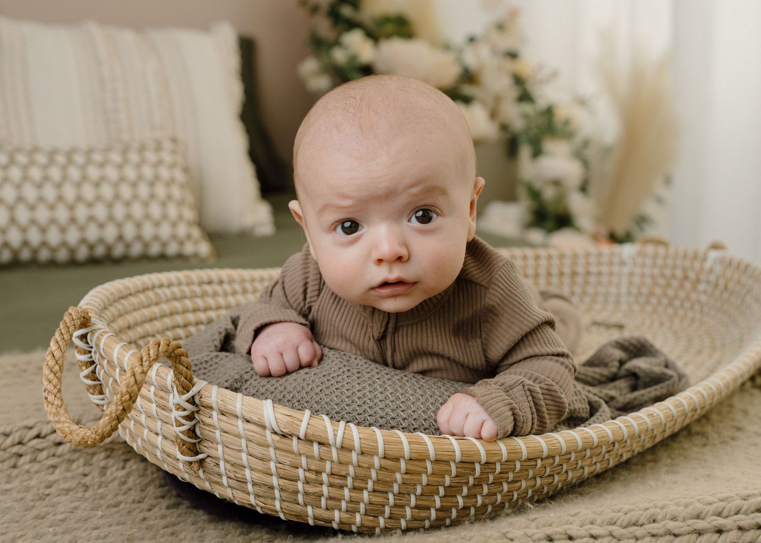 Adorable baby in brown onesie doing tummy time in a woven moses basket.