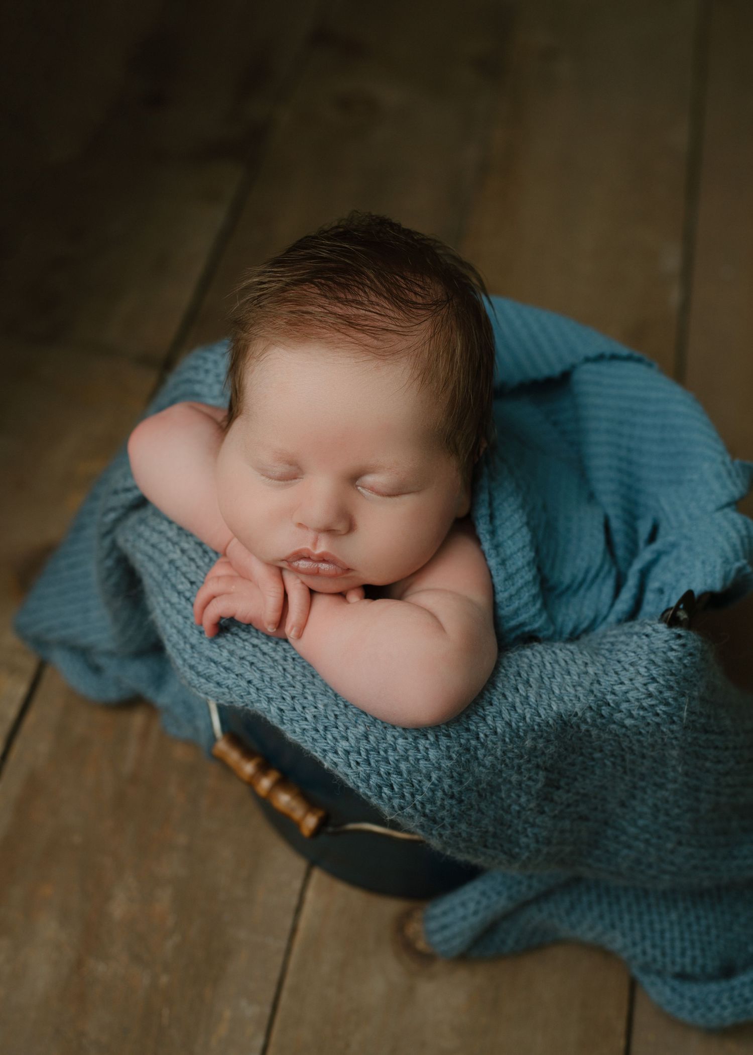 Sleeping newborn baby wrapped in teal knit blanket in a small wooden bowl.