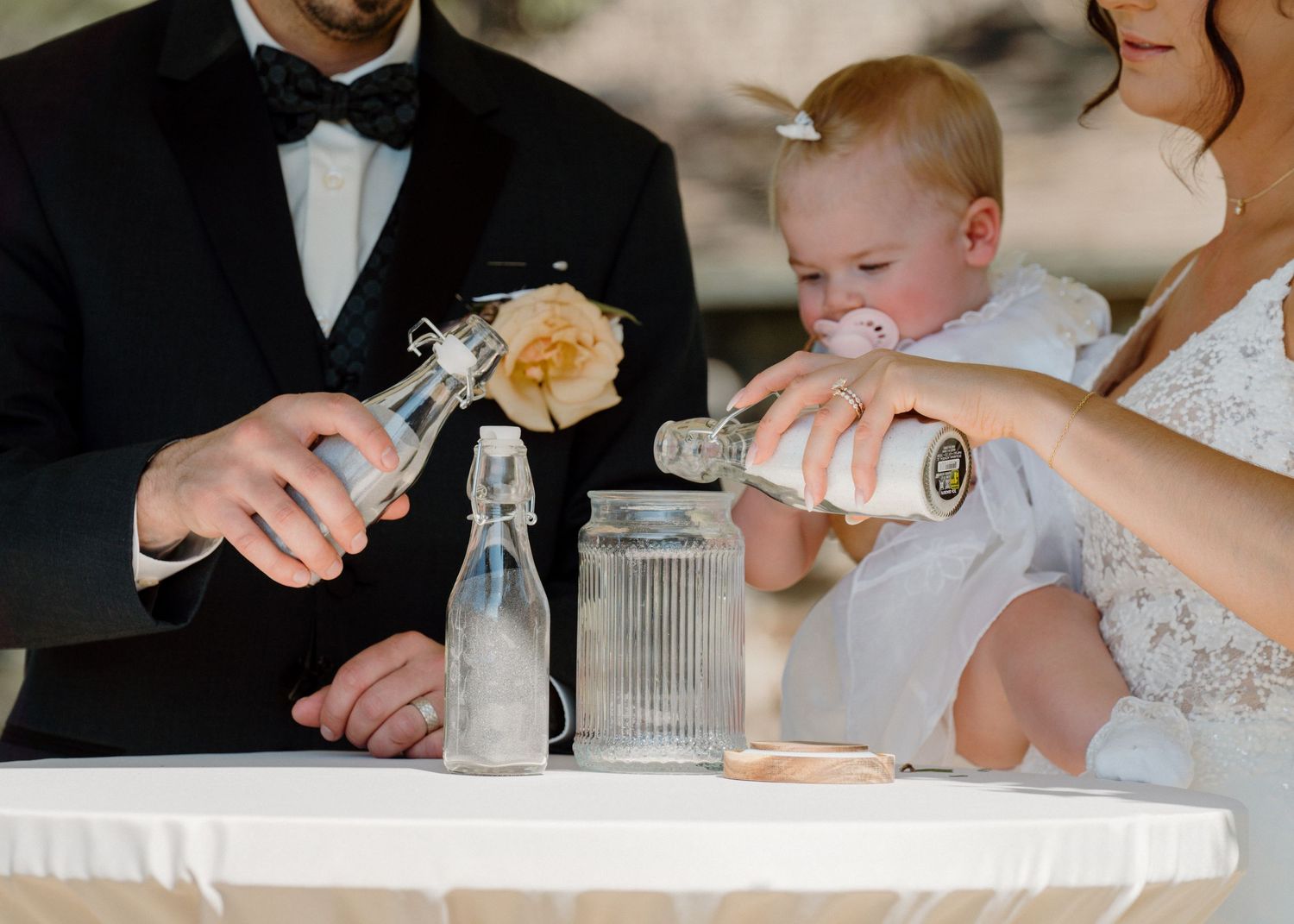 Bride and groom performing a sand unity ceremony while holding their baby in white at a wedding reception table.