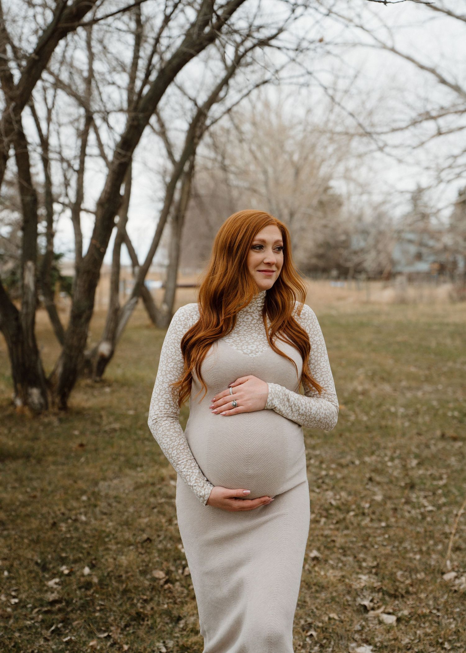 Pregnant woman with long red hair in beige maternity dress holding belly outdoors.