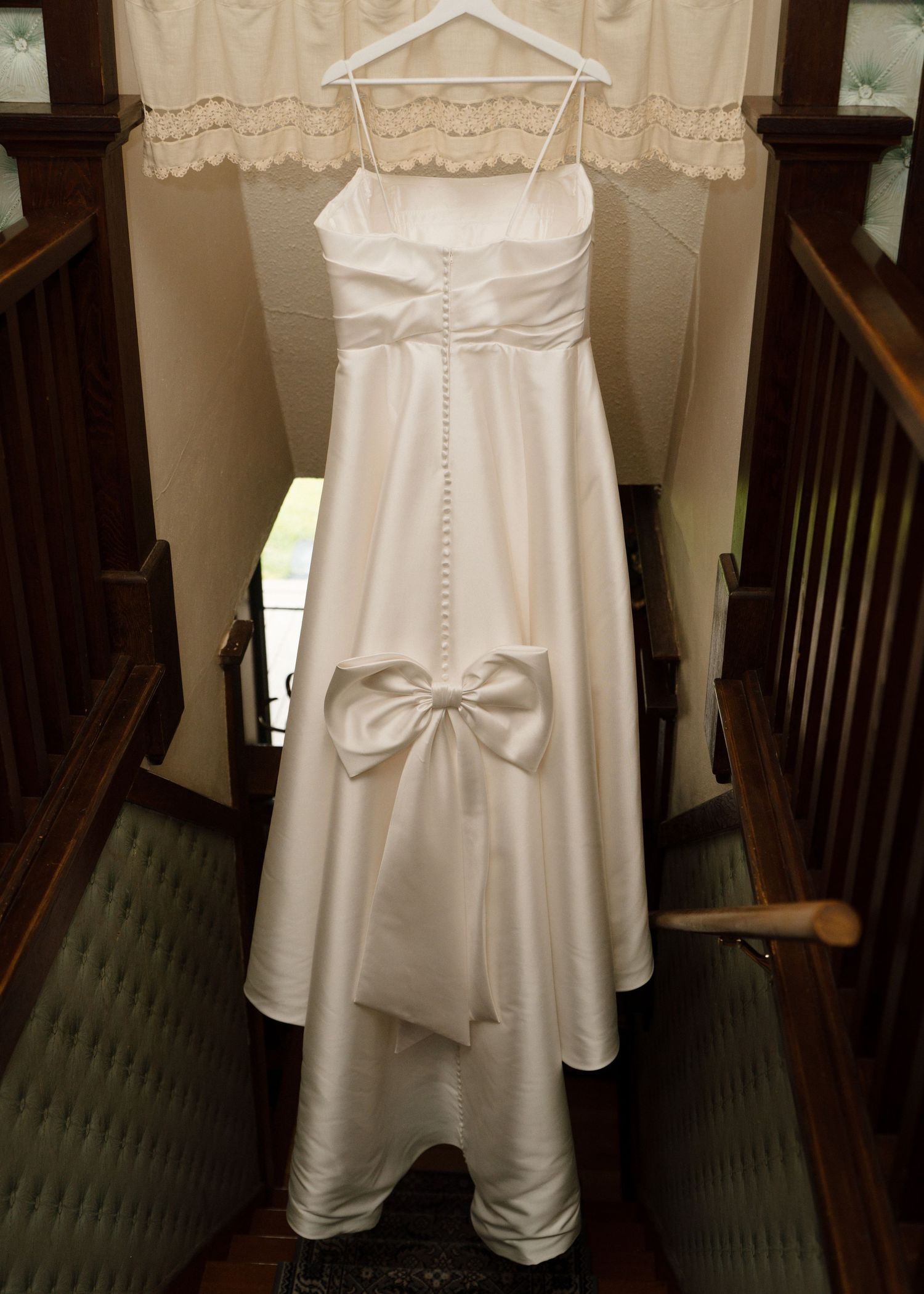Elegant white satin wedding gown with bow detail hanging on a staircase banister.