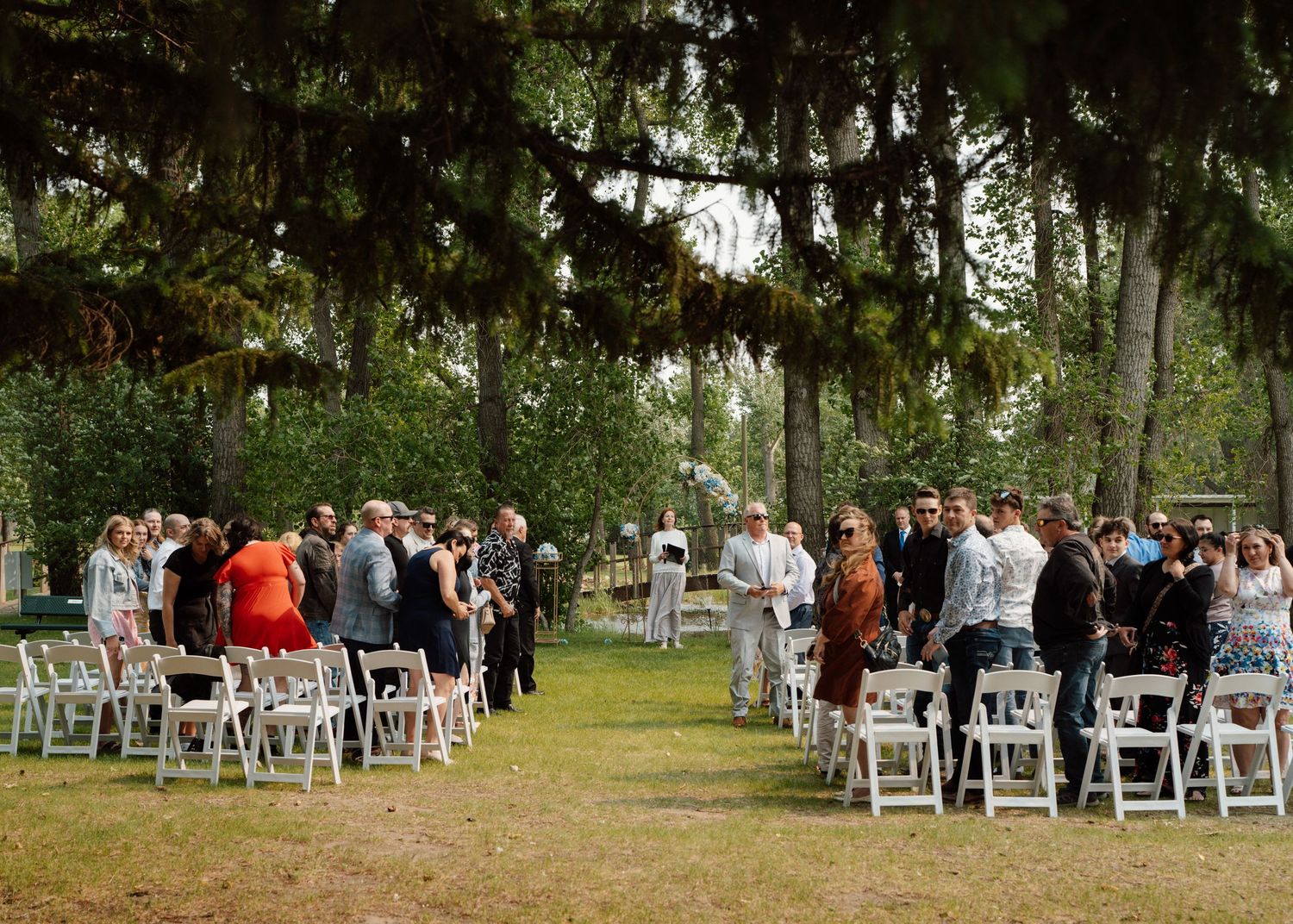 Guests gather at an outdoor wedding ceremony with white chairs under large shady trees.