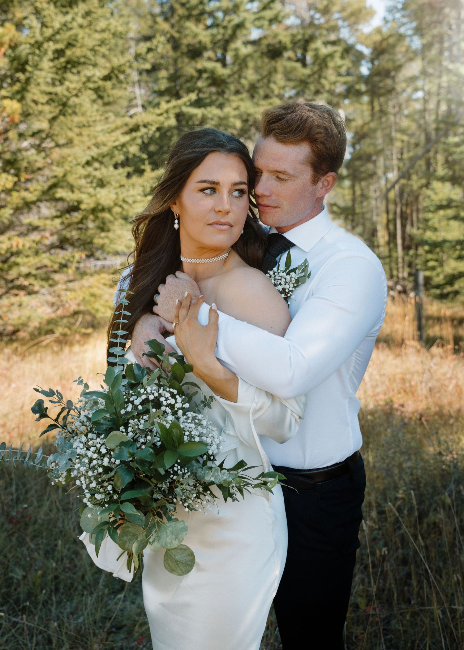 Bride and groom pose intimately in autumn forest meadow, bride holding lush greenery bouquet.