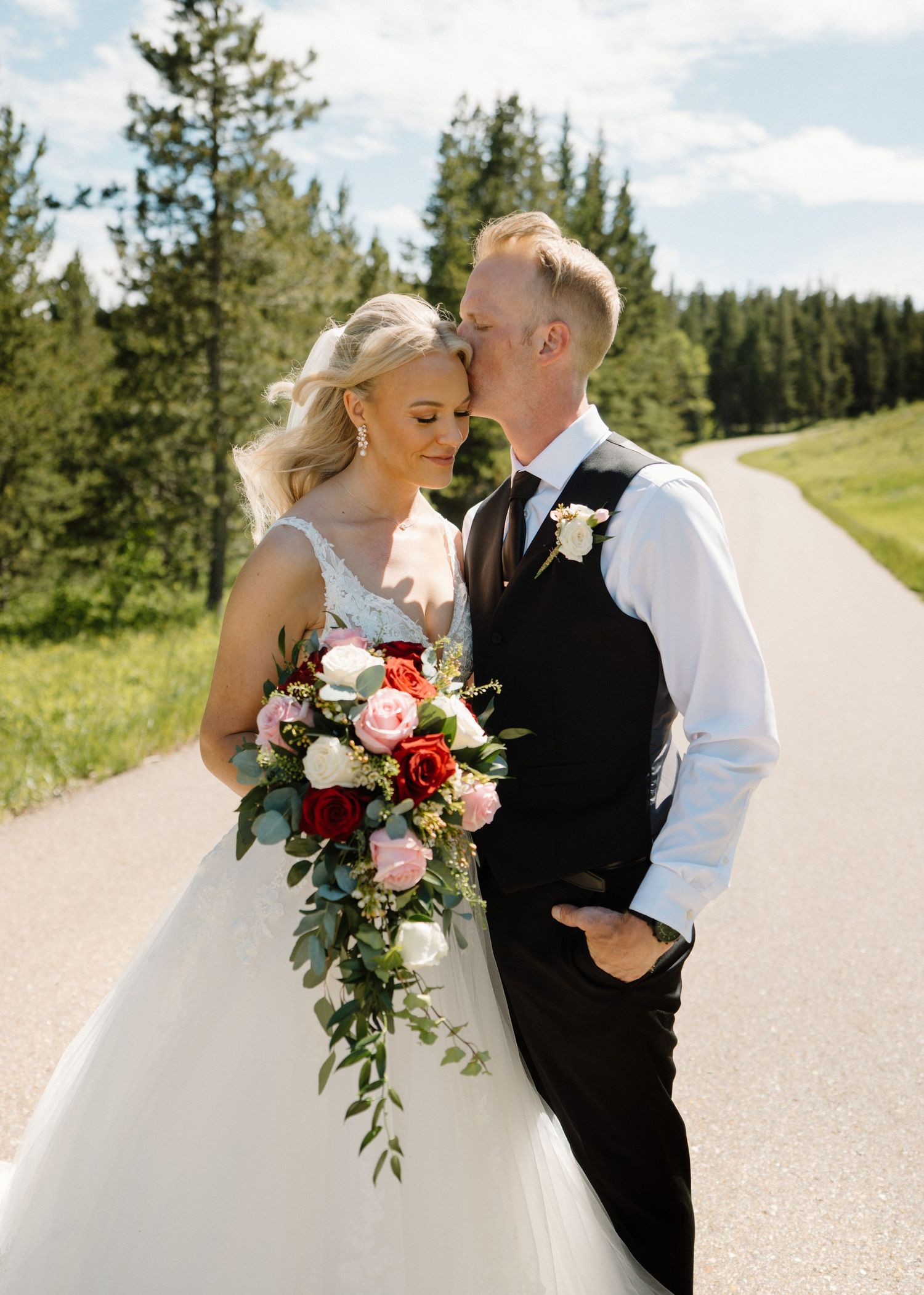 Bride and groom share a romantic moment on a sunlit road surrounded by evergreen trees, bride holding a red and pink bouquet.