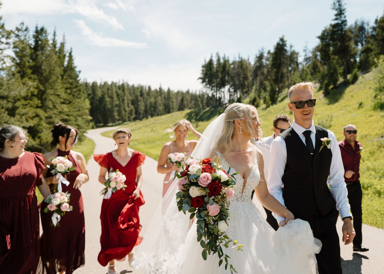 Bride and groom walk with wedding party on a scenic mountain path, bridesmaids in burgundy dresses carrying bouquets.