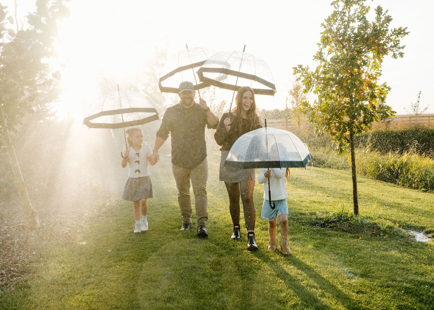 Family of four walking in sunny garden with clear umbrellas during light rain, surrounded by lush green trees.