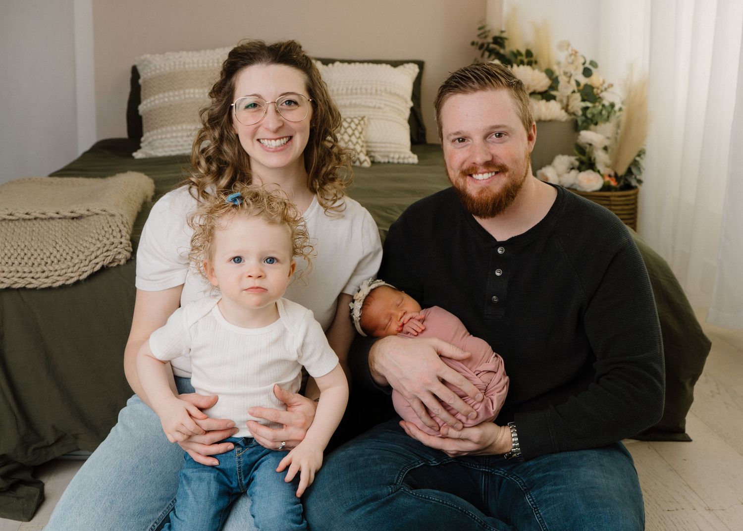 Smiling family of four with toddler and father holding newborn baby posed in front of a cozy bedroom backdrop.
