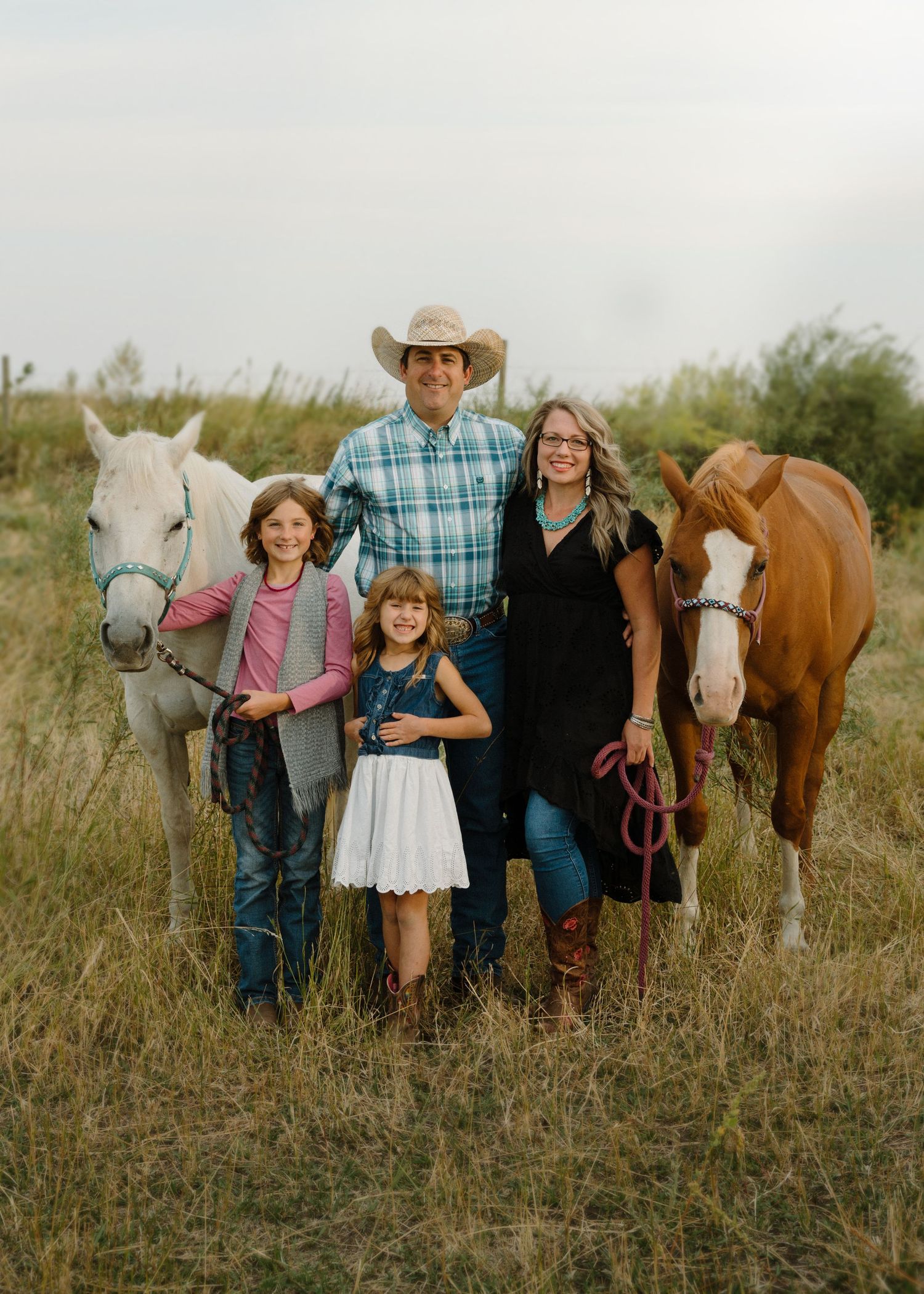 Family with two girls and two horses posing in field with father wearing cowboy hat.