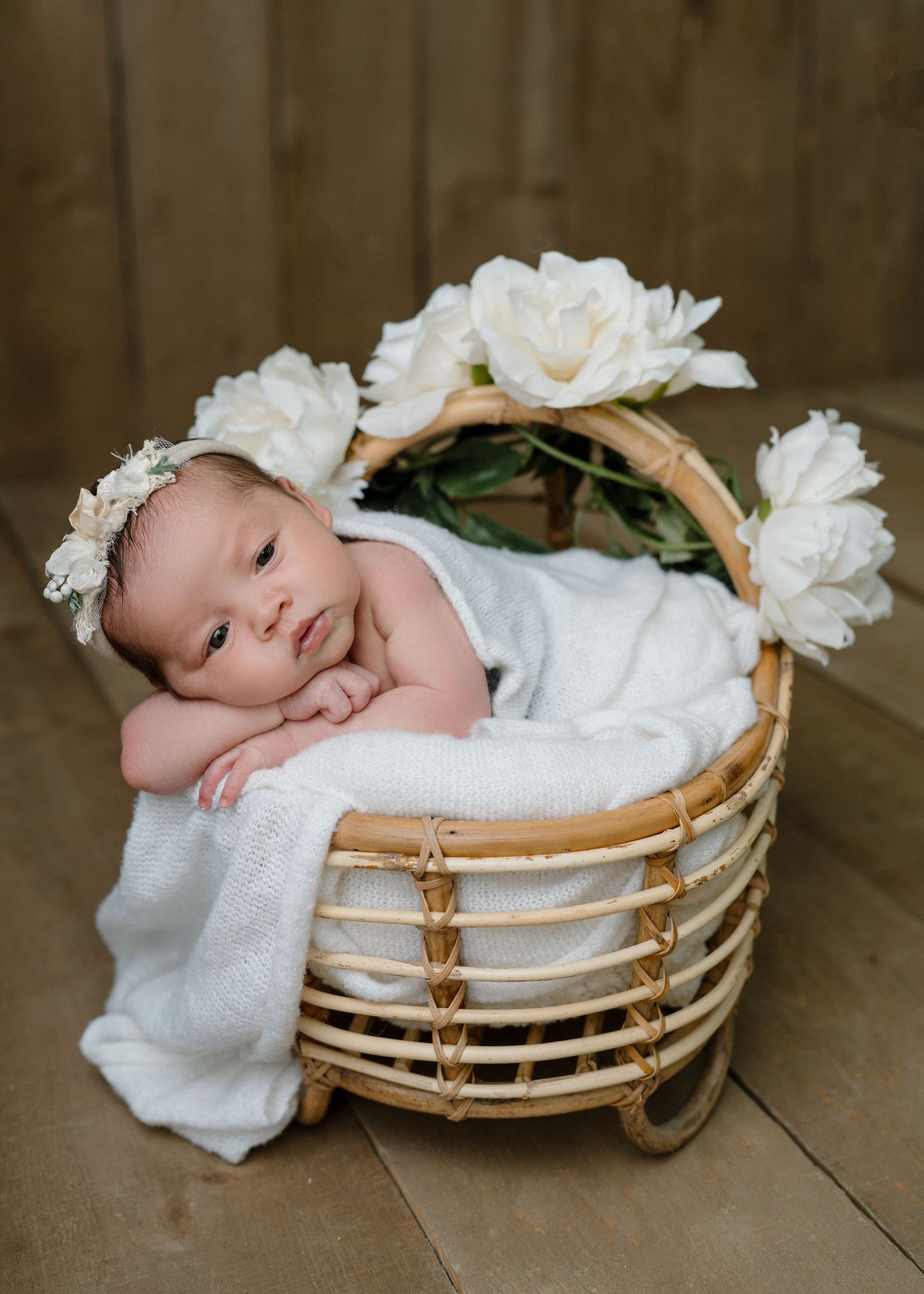 Alert newborn baby girl resting in wicker basket with white flowers and floral headband on wood floor.