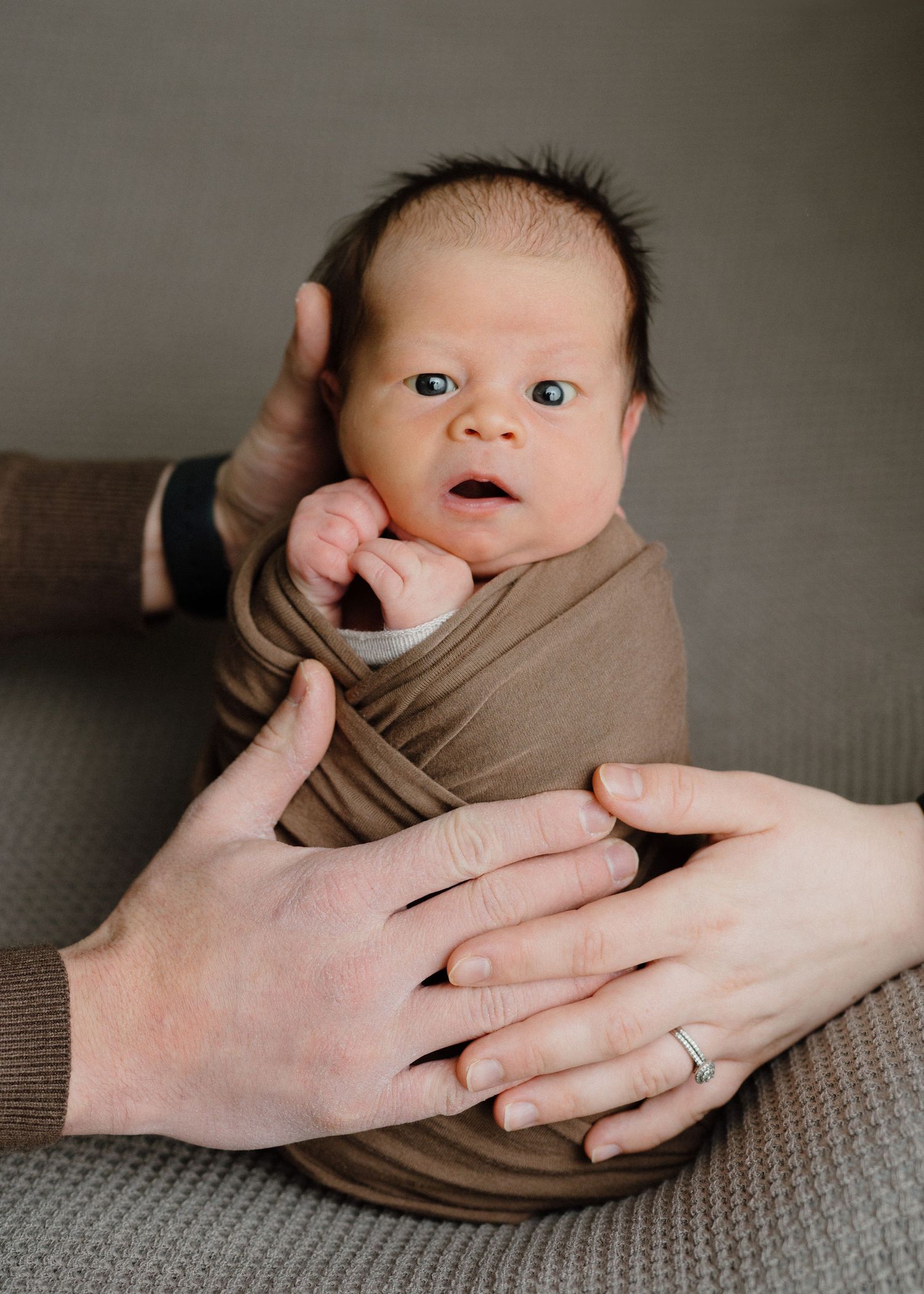Newborn baby swaddled in brown wrap held by two parents hands, one wearing a wedding ring.