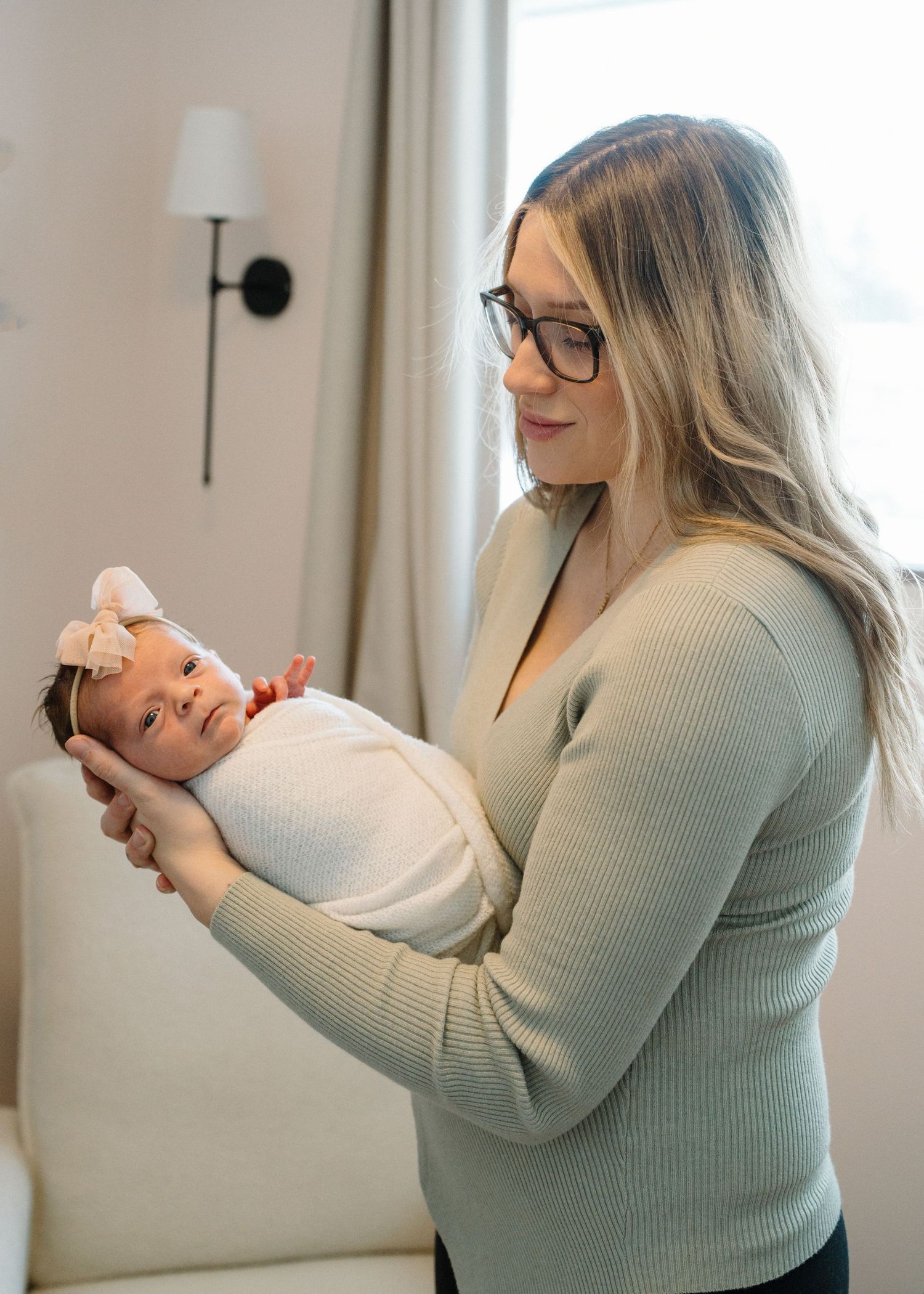 Blonde mother wearing glasses holding swaddled newborn with pink bow headband by window.