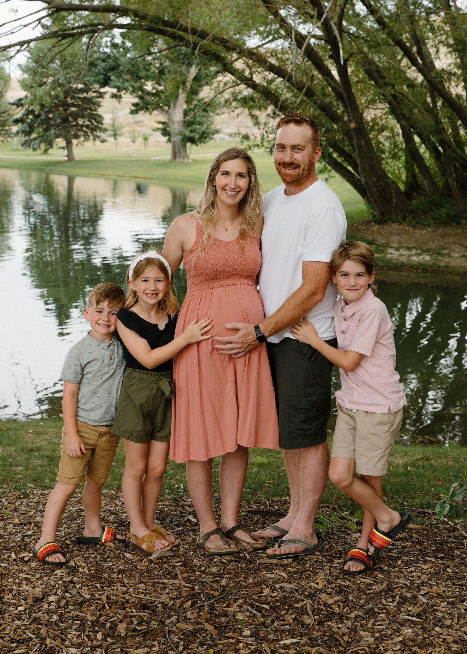 Happy family of five with pregnant mother in coral dress posing by a pond.