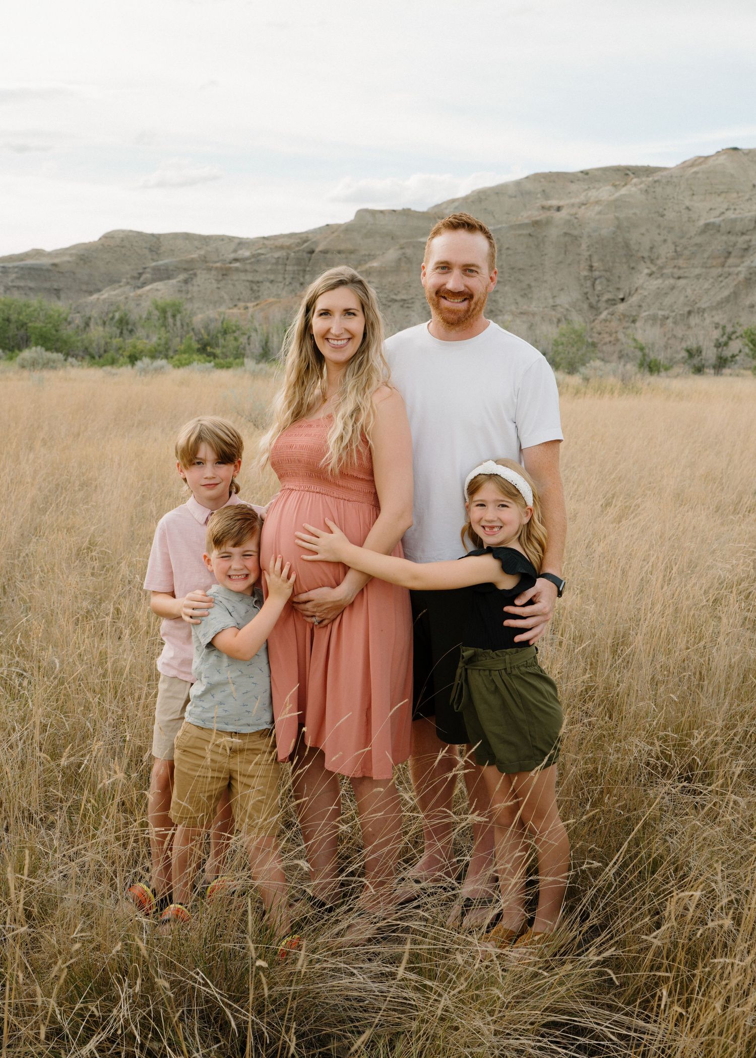Family of five posing in a golden field with hills, pregnant mother in coral dress.