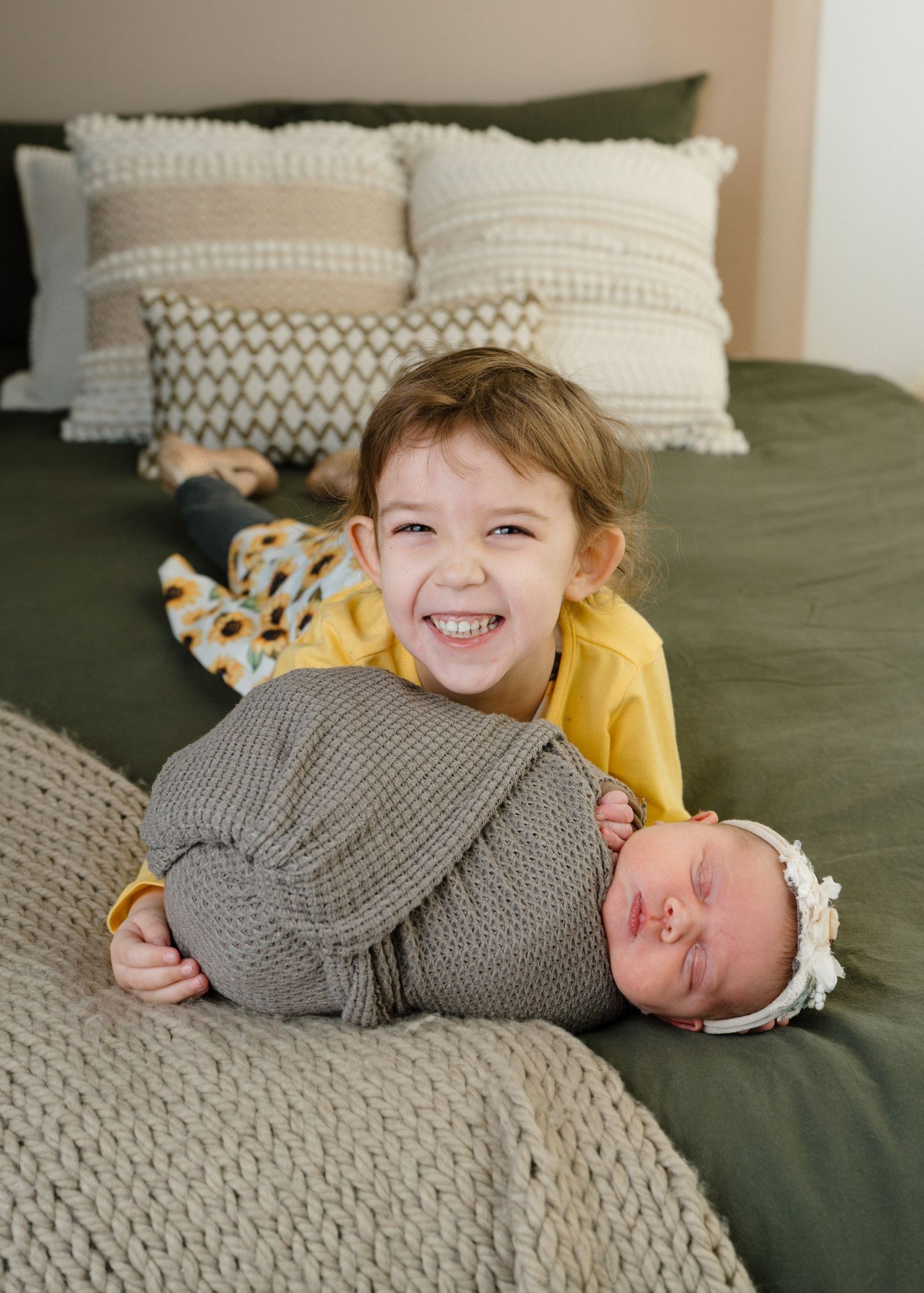 Smiling toddler in yellow shirt proudly holds sleeping swaddled newborn sibling on green bed.