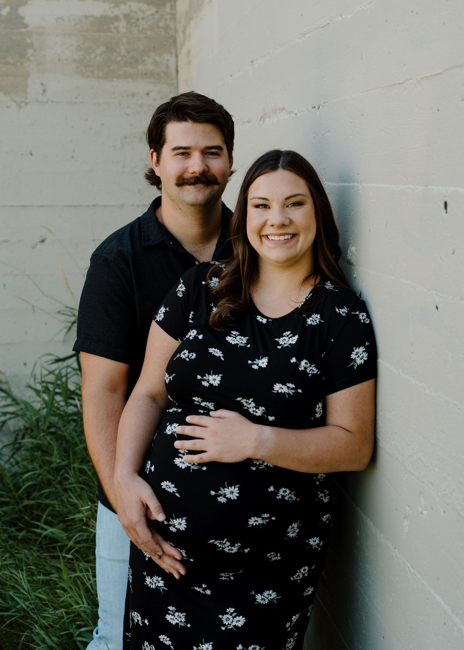Pregnant couple in matching black outfits smile together against a concrete wall outdoors.