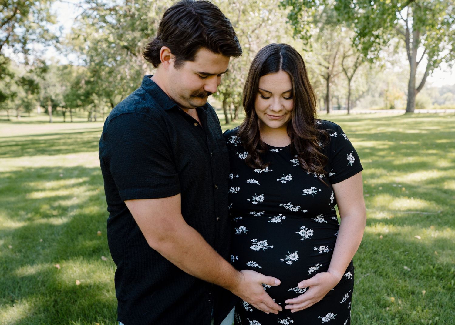 Expecting couple looks down lovingly at baby bump while standing in a sunny green park.