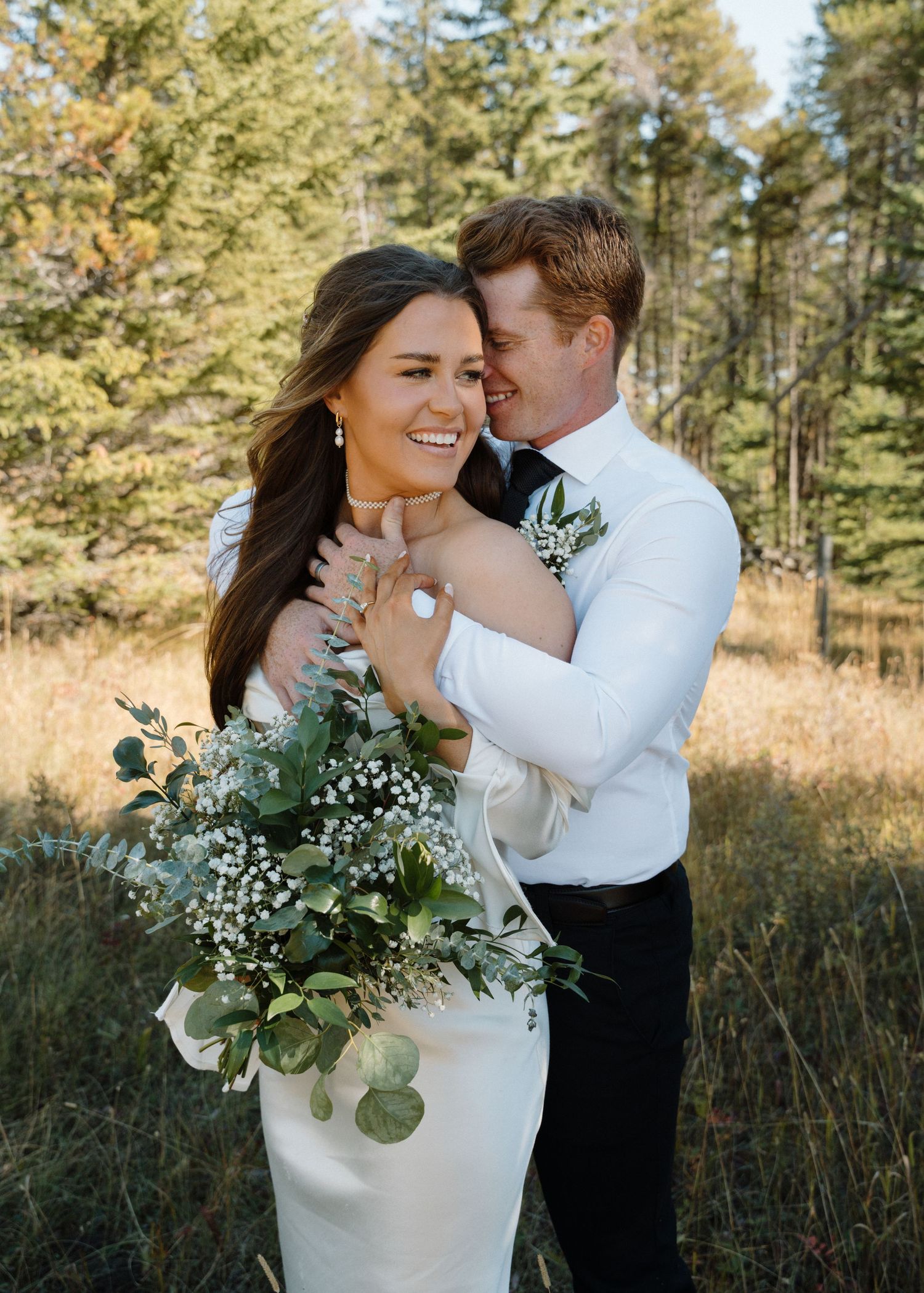 Groom embraces smiling bride holding greenery bouquet in a sunlit forest meadow setting.