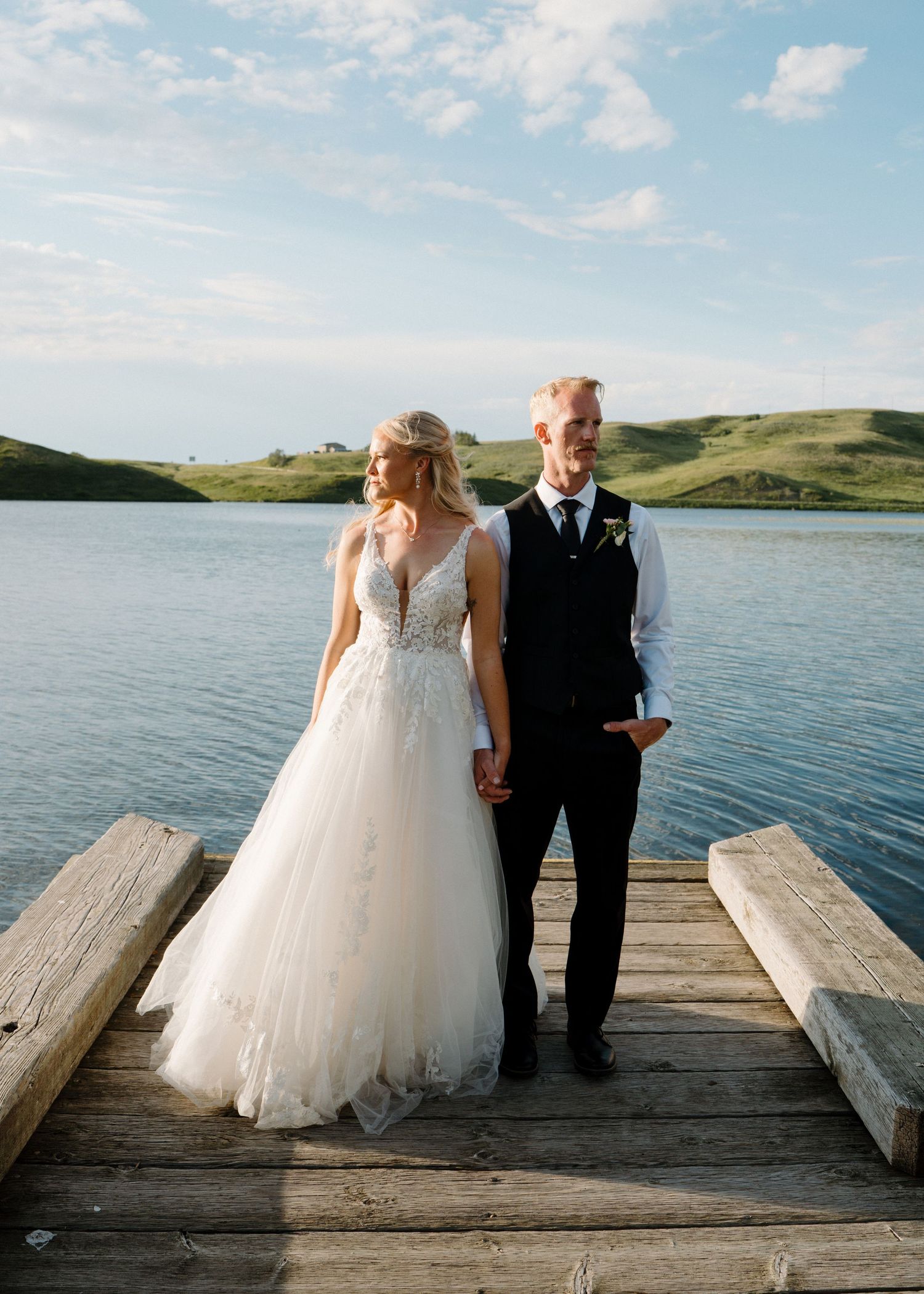 A bride and groom hold hands on a wooden dock overlooking a serene lake with green hills under a blue sky.