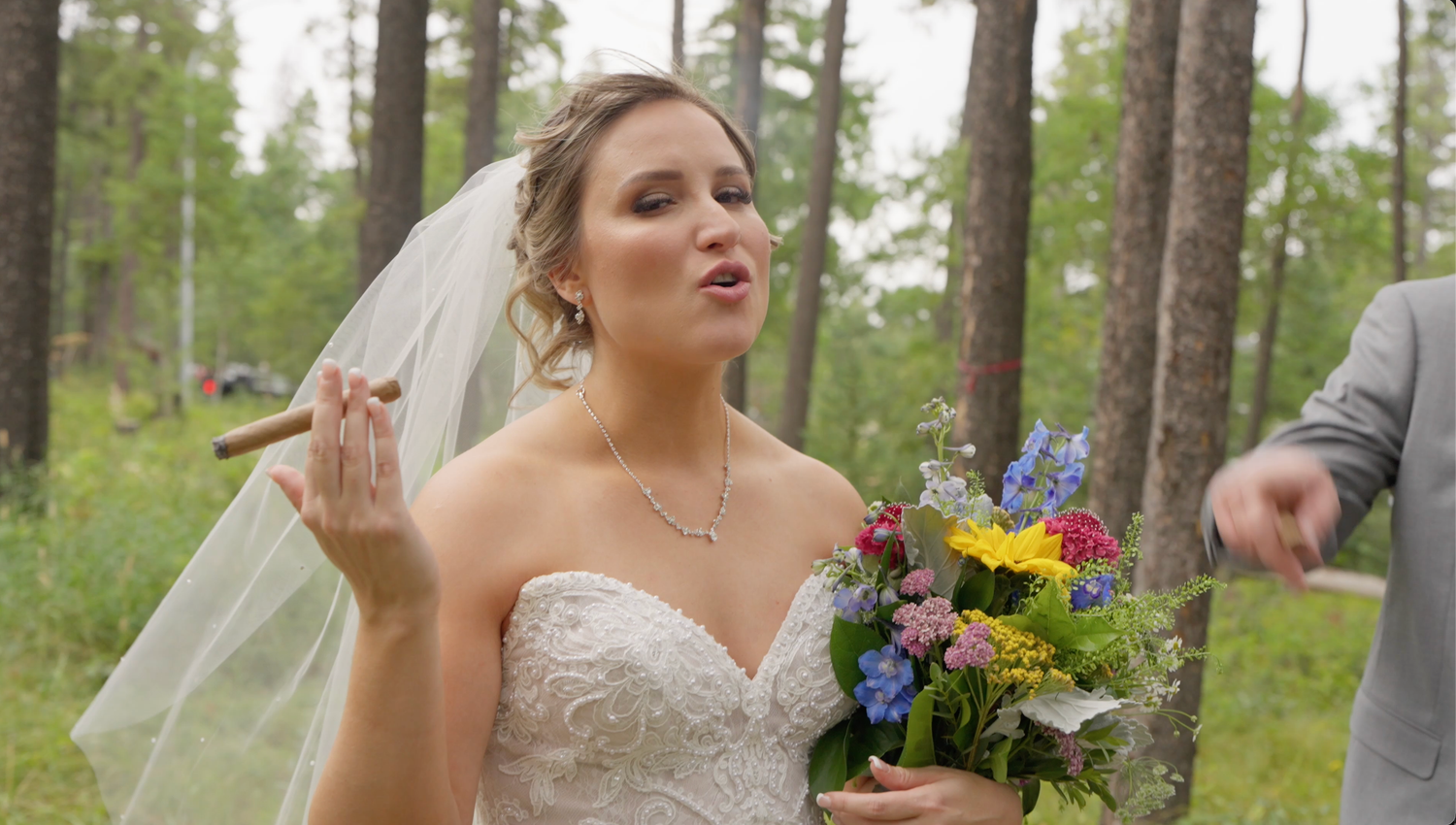 Bride in white lace gown holding colorful bouquet and cigar, posing outdoors in forest setting on wedding day.