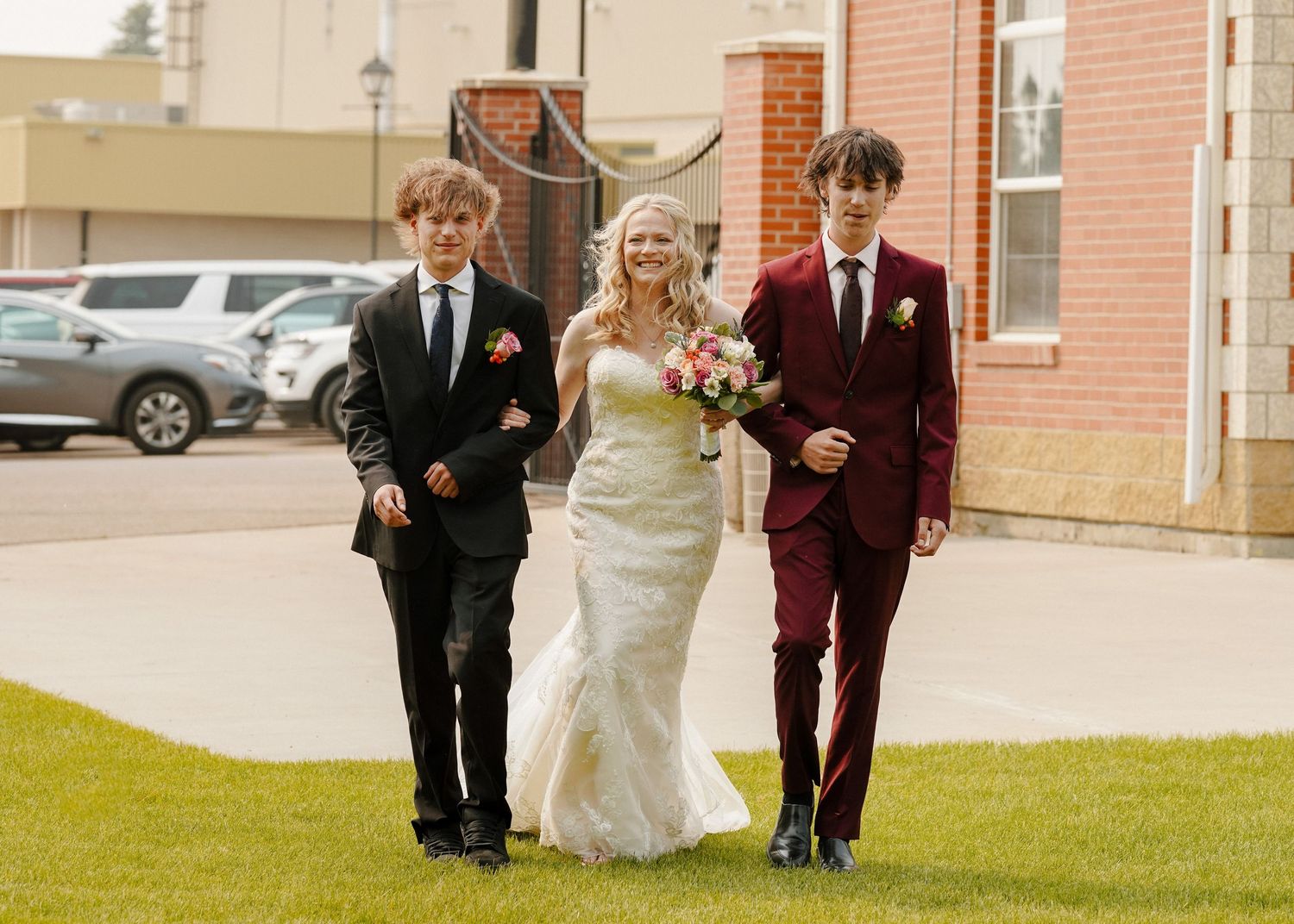 Bride in white lace gown walks with two young men in suits on a sunny outdoor wedding day.