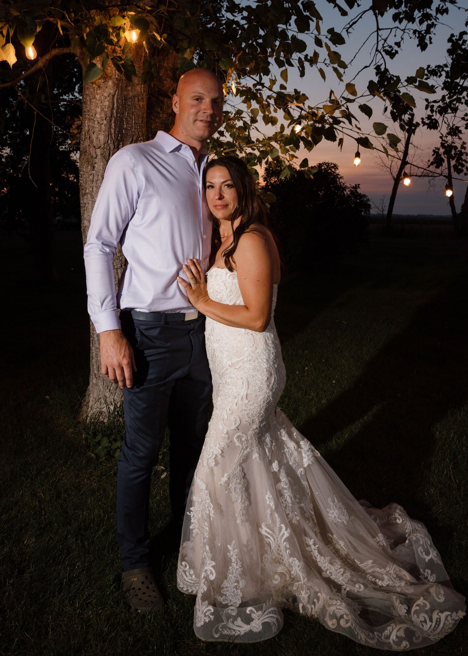 Elegant bride in lace mermaid gown and tall groom pose under tree with string lights at twilight.