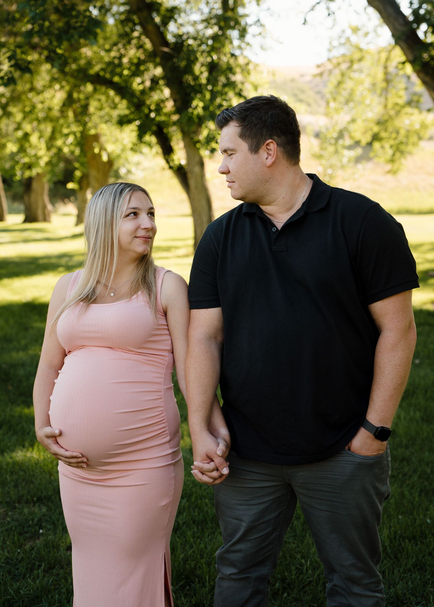 Expecting couple holding hands in a sunny park, woman in pink maternity dress cradling her baby bump.