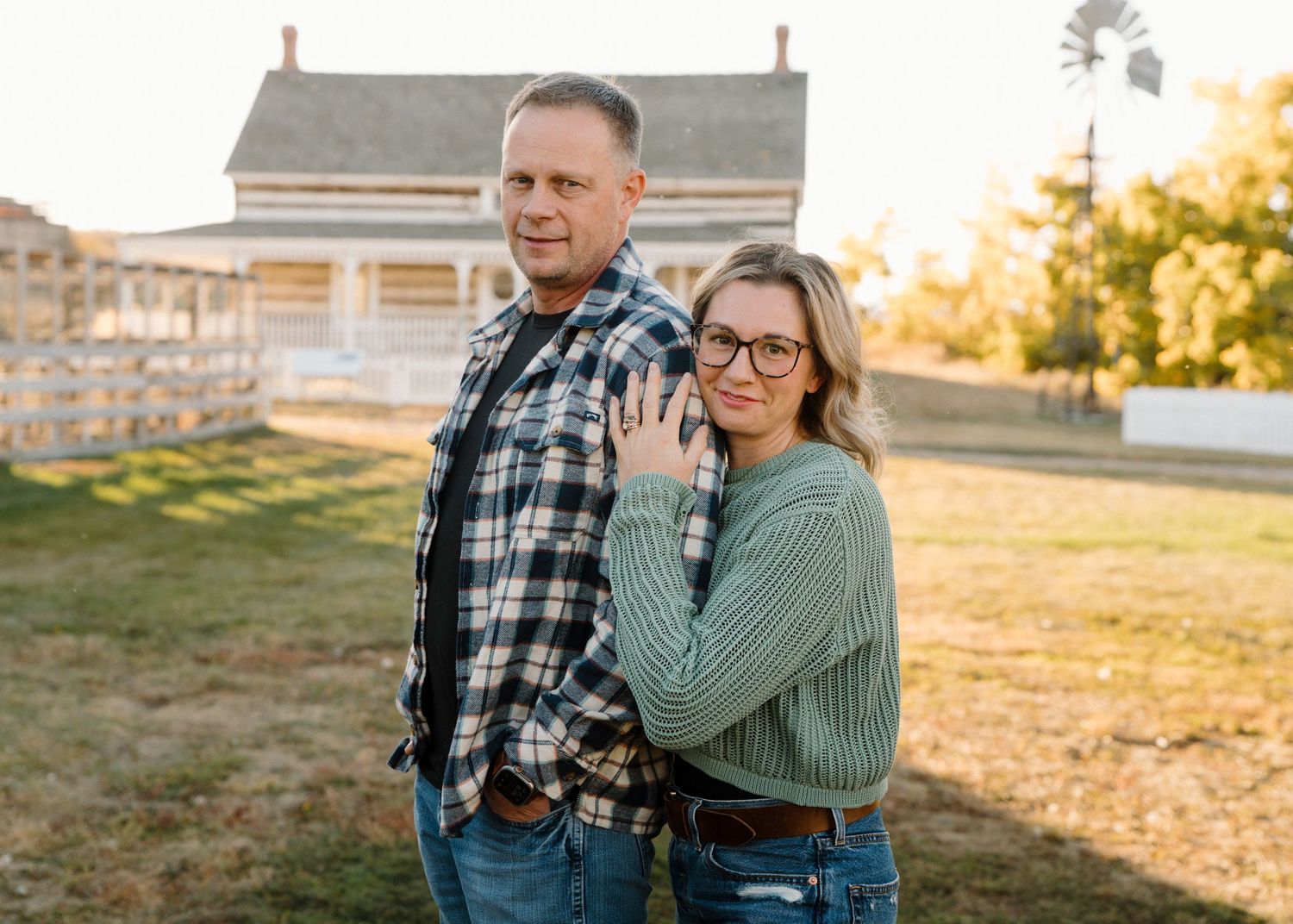 Couple poses together in front of a farmhouse at sunset, woman resting hand on mans back.