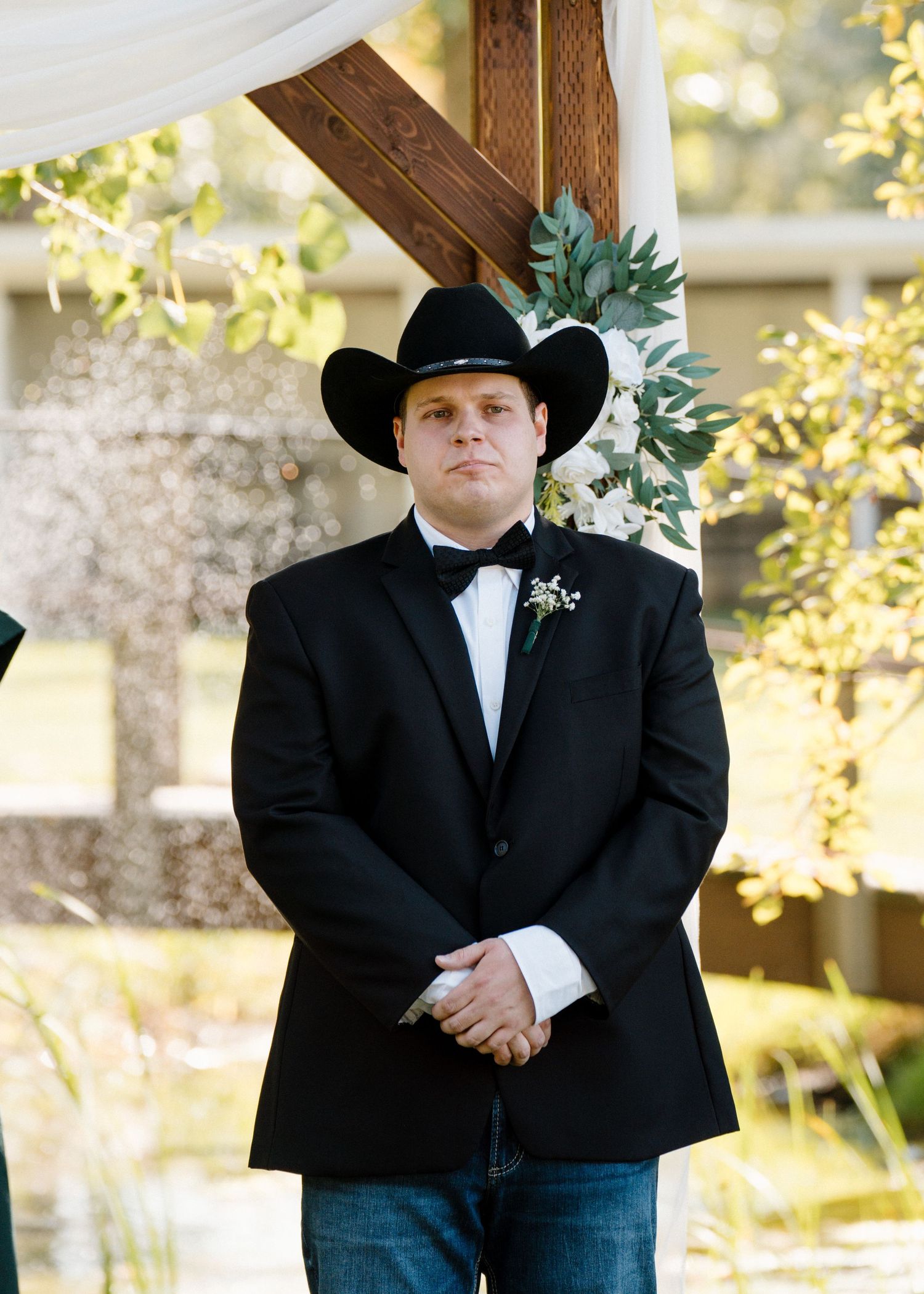 Groom in black tuxedo and cowboy hat stands at wedding altar with boutonniere and bow tie.