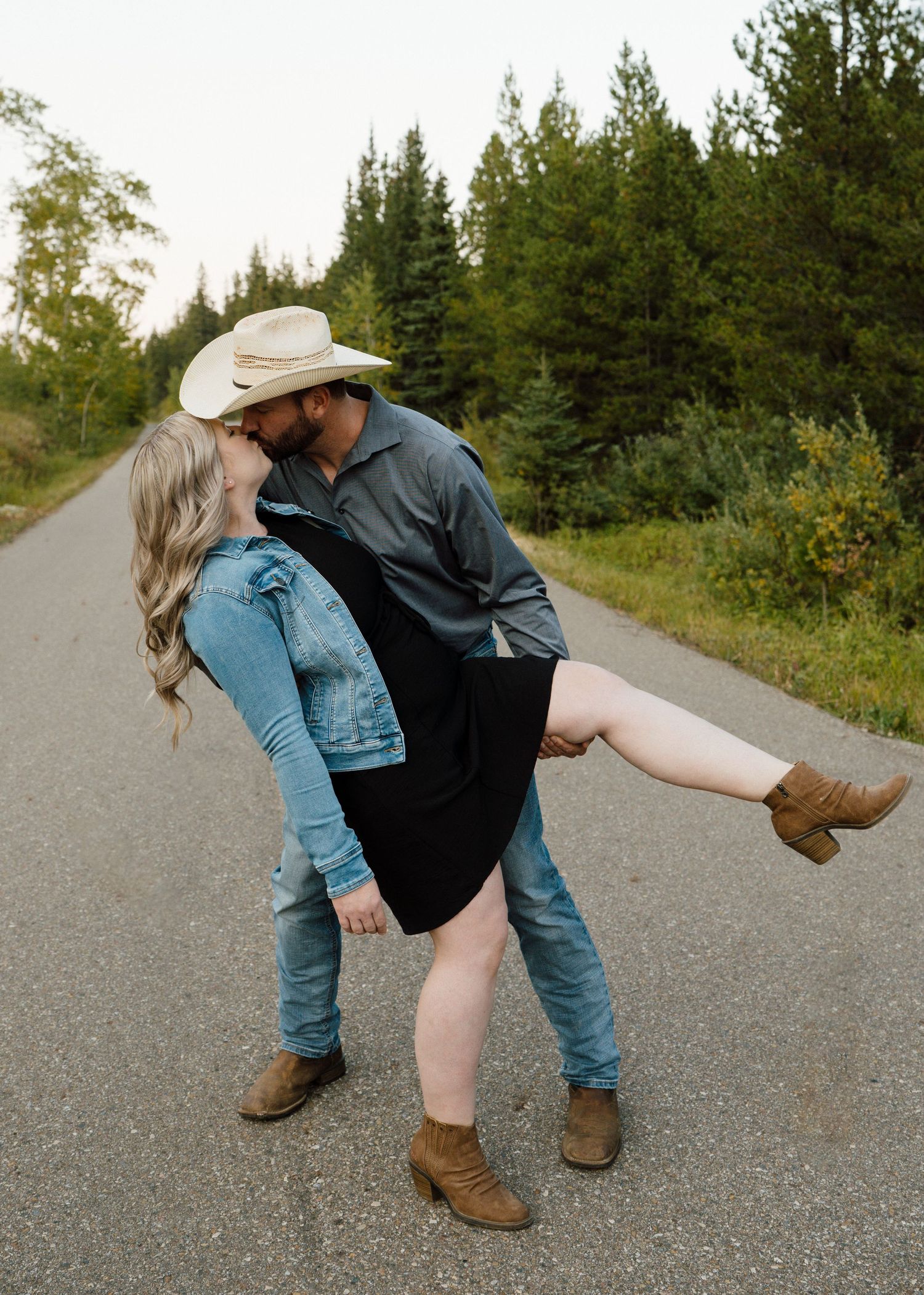 Man in cowboy hat dips woman in black dress on rural road lined with green trees.