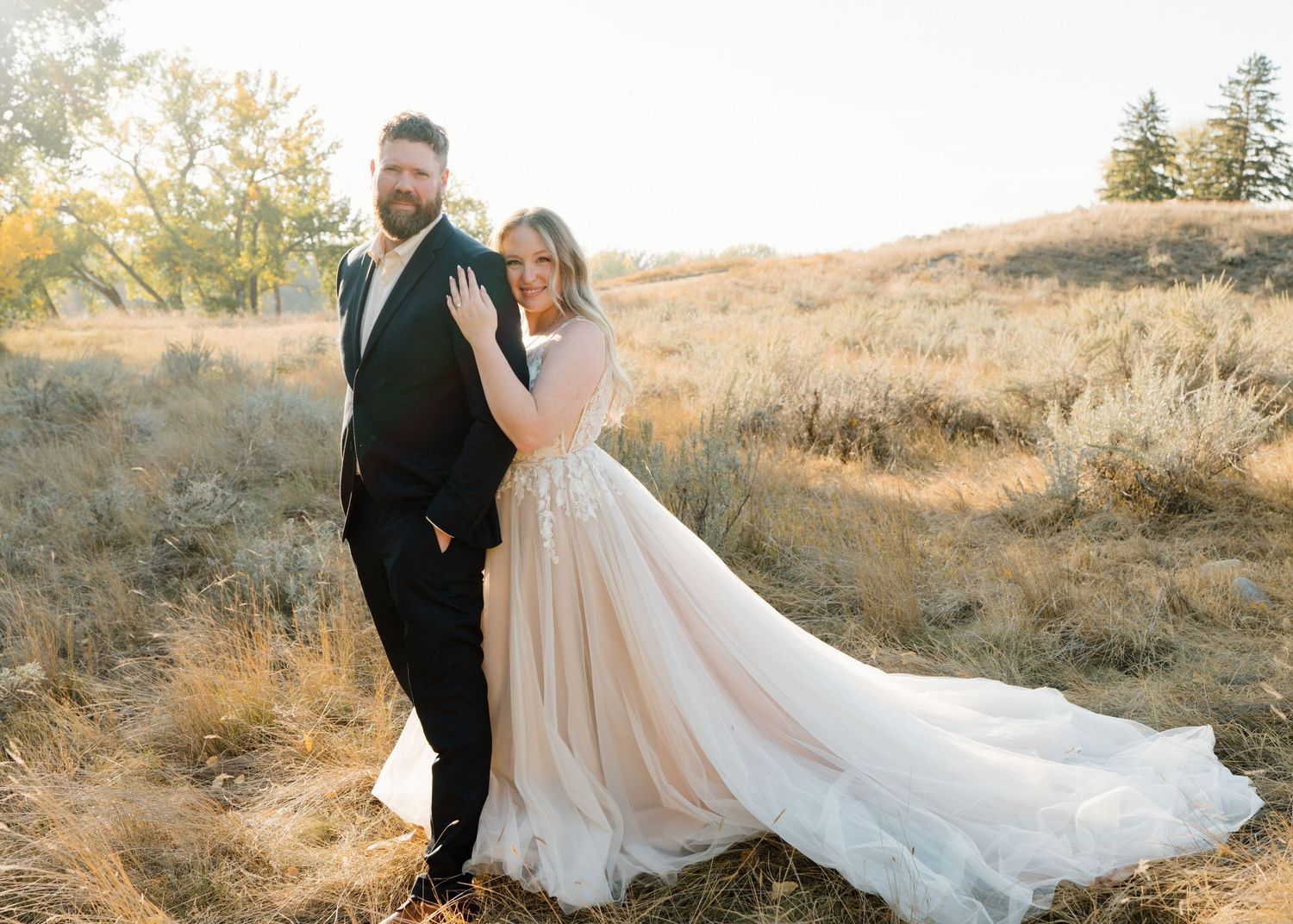 Bride in flowing blush wedding gown and bearded groom posing in golden autumn field at sunset.