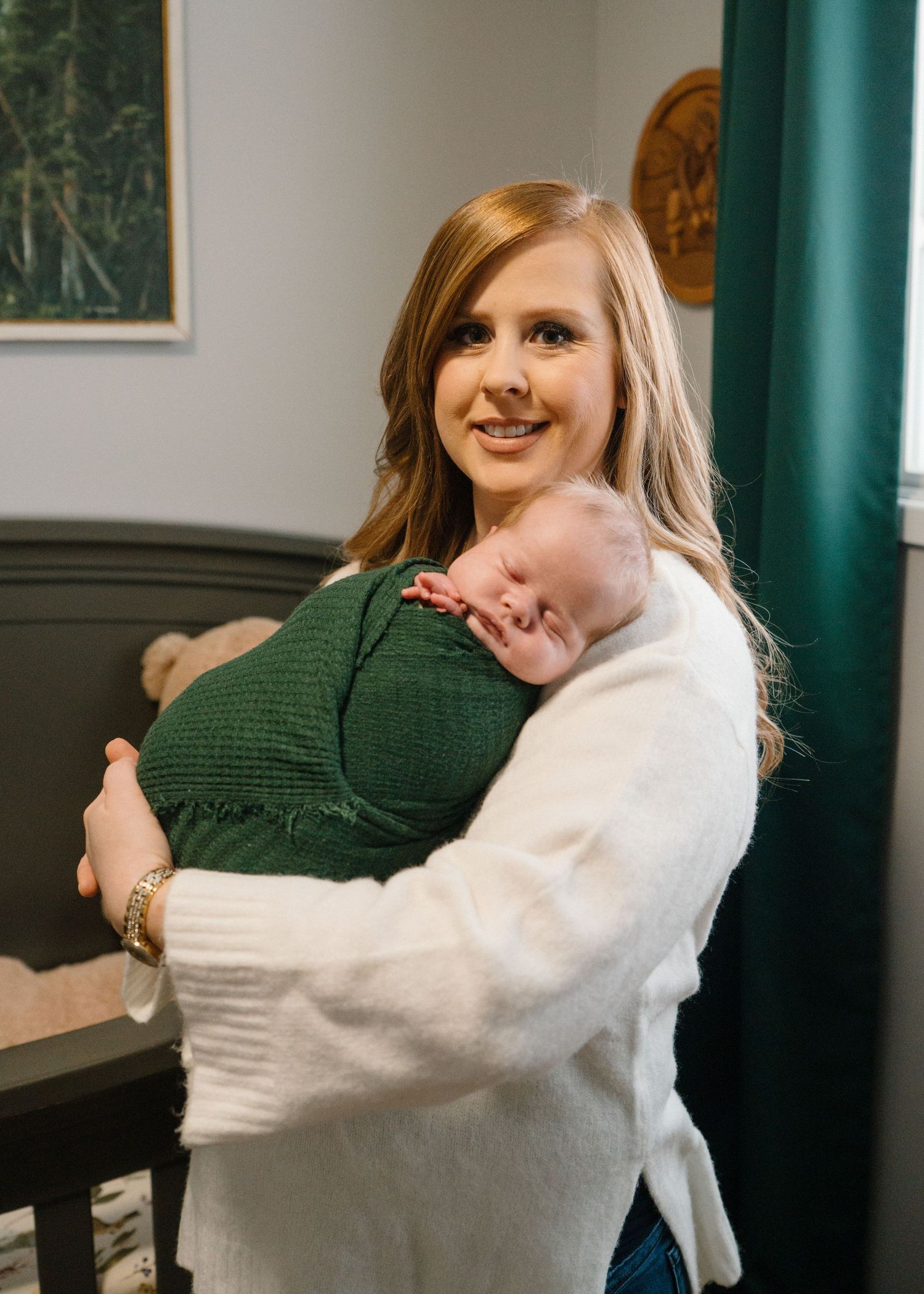 Smiling red-haired mother in white sweater holding sleeping newborn baby against her chest.