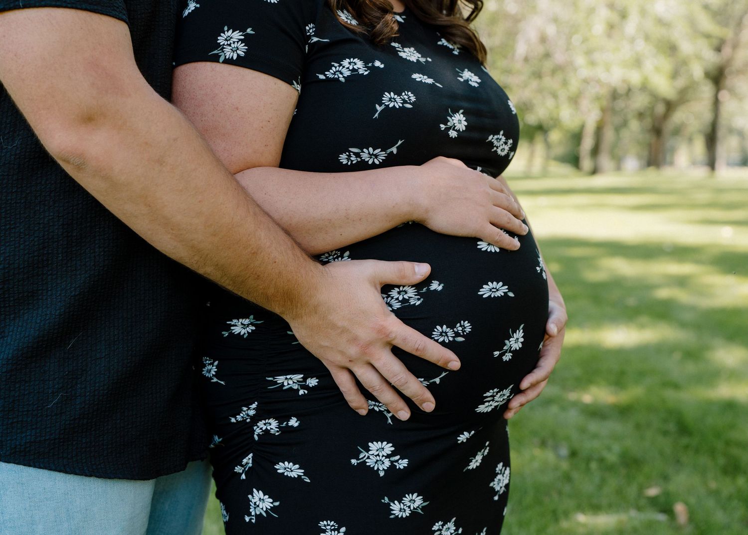 Couple cradles pregnant belly together outdoors in a lush green park setting.